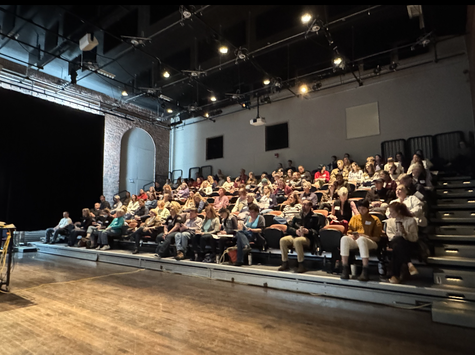 Audience seated in a small theater or lecture hall, waiting for a presentation or performance to begin.