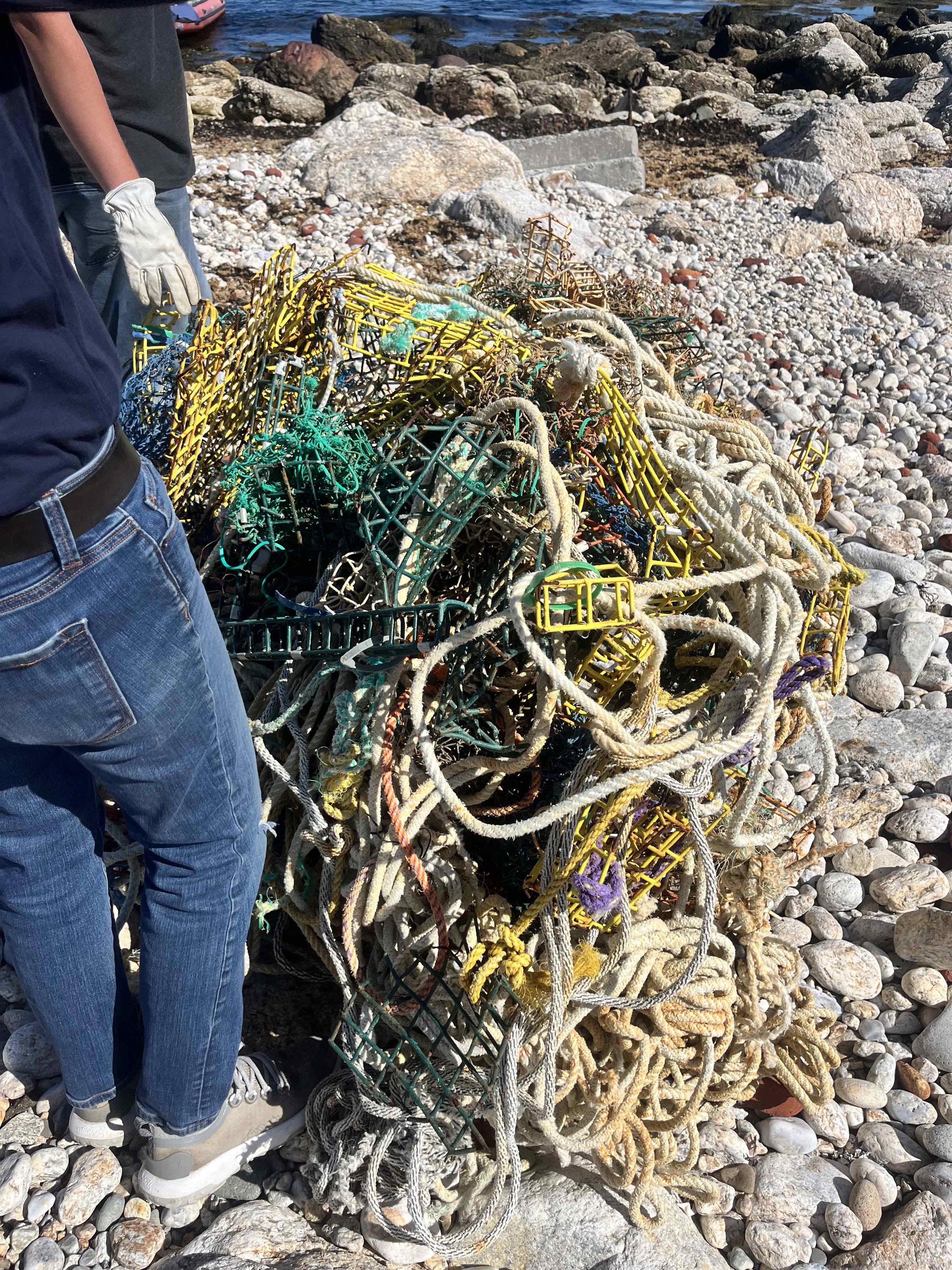 A pile of tangled lobster traps and ropes on a rocky beach with the sea in the background.