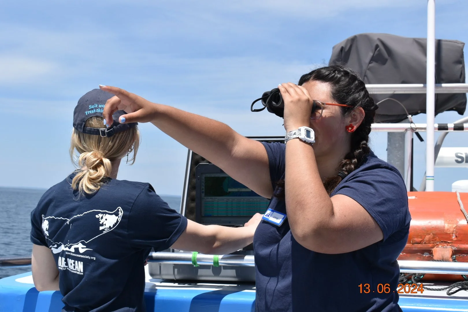 Two women on a boat, one looking through binoculars and the other working on a laptop, both wearing navy blue shirts with a whale logo, the ocean in the background, and a blue sky with some clouds.