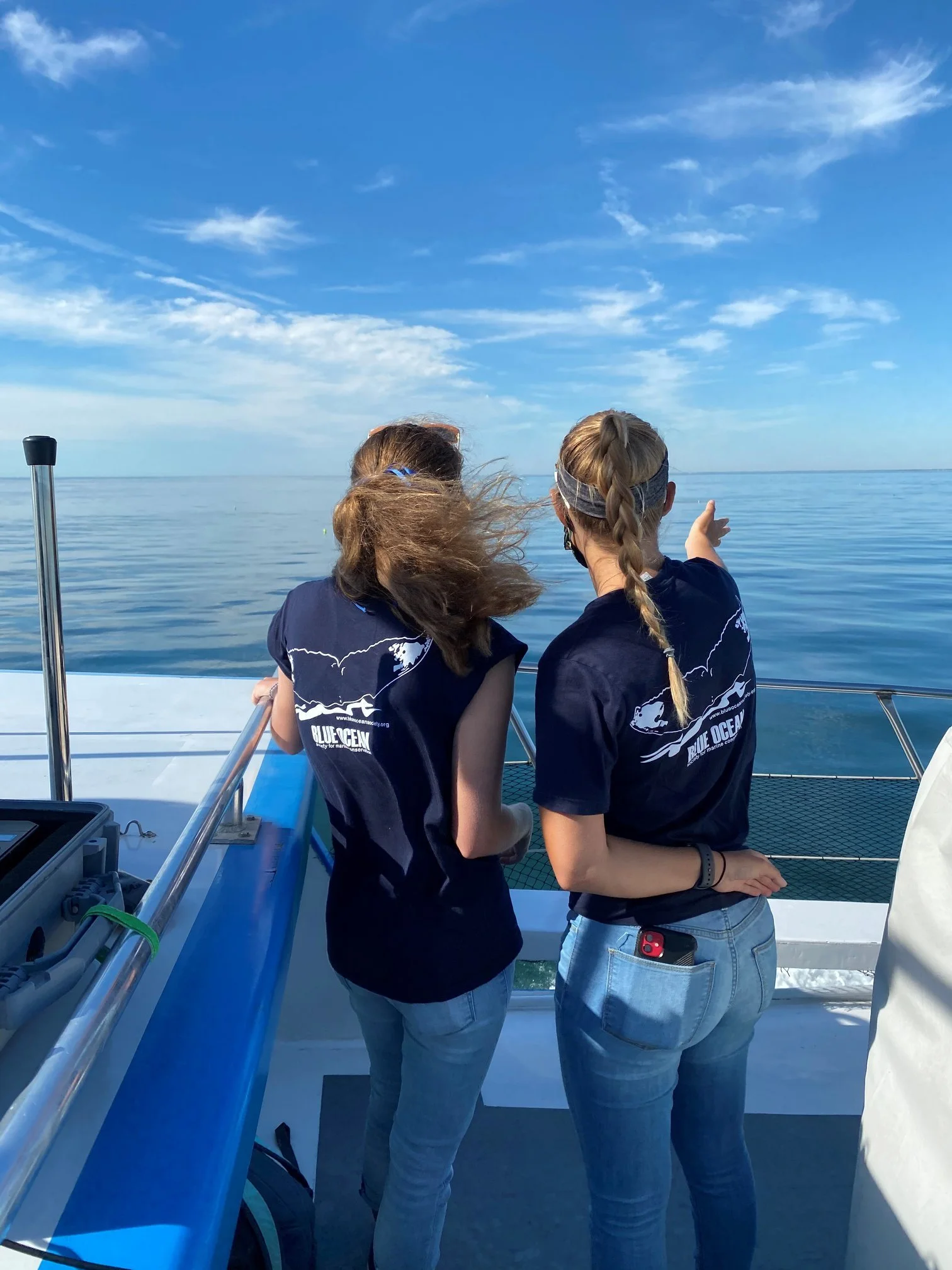 Two women on a boat, looking out over a calm ocean with a blue sky and some clouds, during daytime.