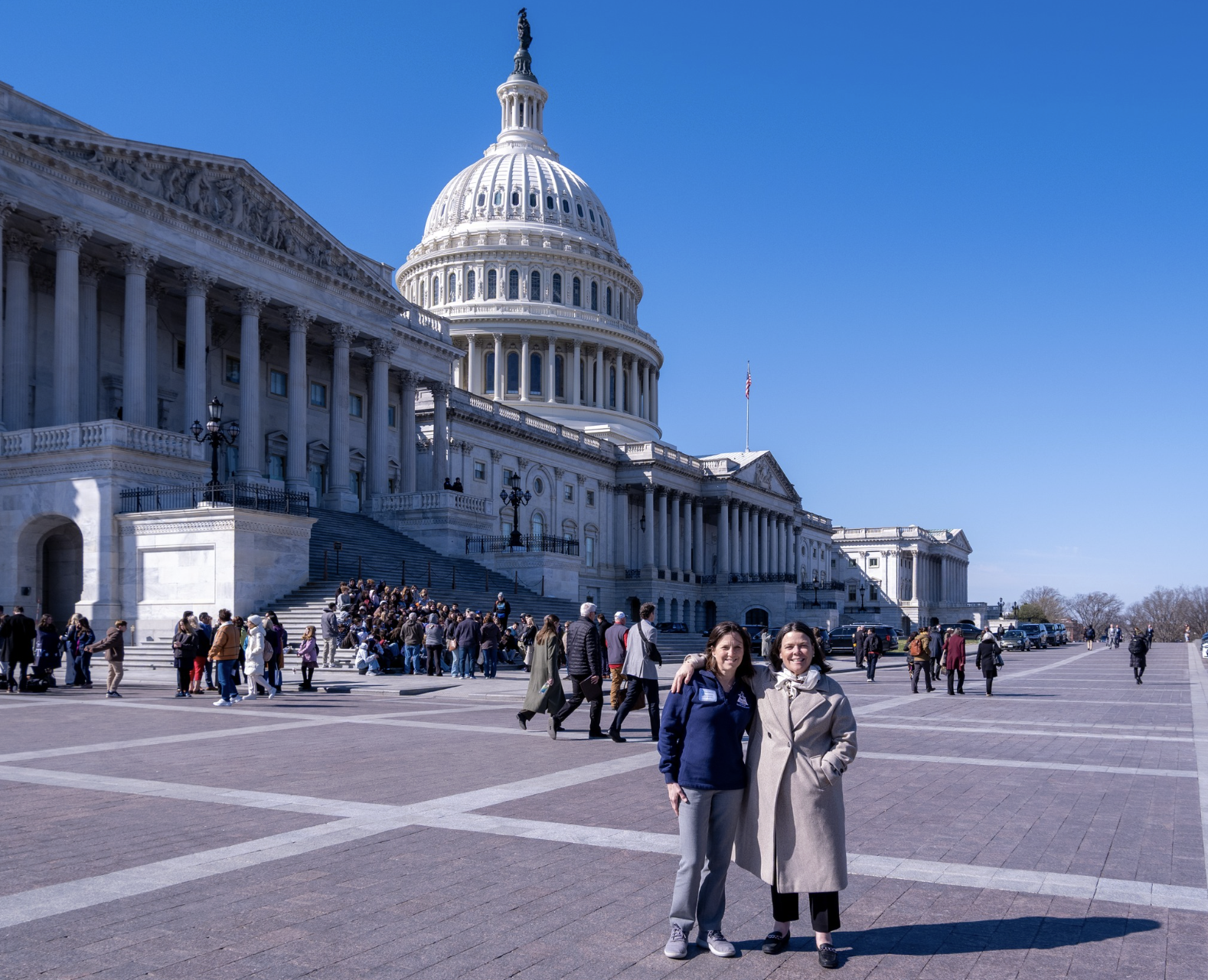The United States Capitol building in Washington, D.C., with two women smiling at the camera in the foreground. Many visitors are gathered on the steps and around the area on a sunny day.