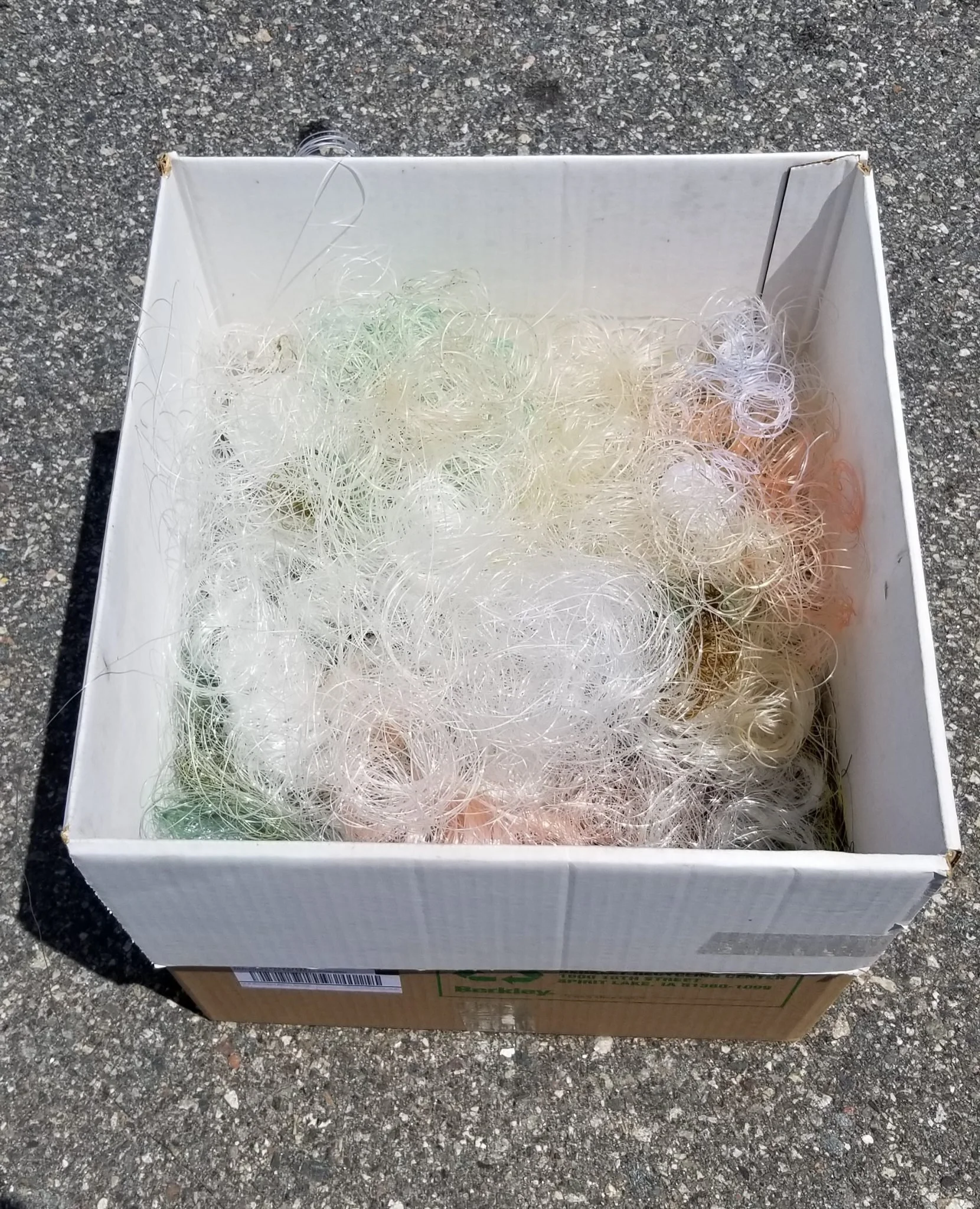 A rectangular cardboard box with a white interior, filled with colorful rubber bands, sitting on an outdoor asphalt surface.