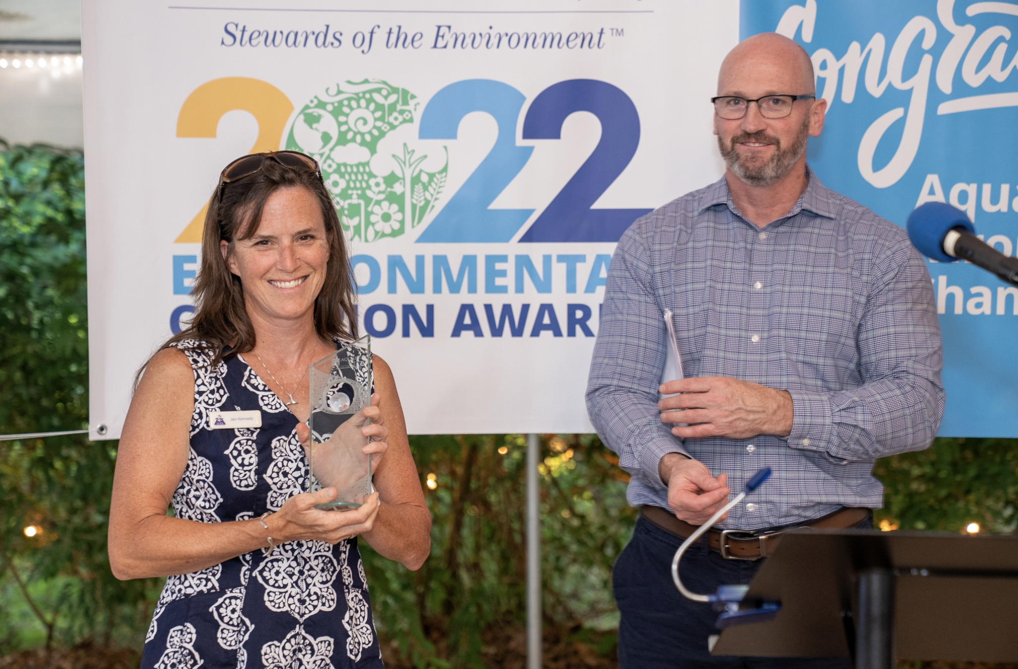 A woman holding a transparent award and smiling, standing next to a man with glasses, in front of a large banner that reads 'Stewards of the Environment, 2022 Environmental Change Award.' They are outdoors, and the woman is wearing a navy and white patterned dress, while the man is wearing glasses and a light purple checked shirt.