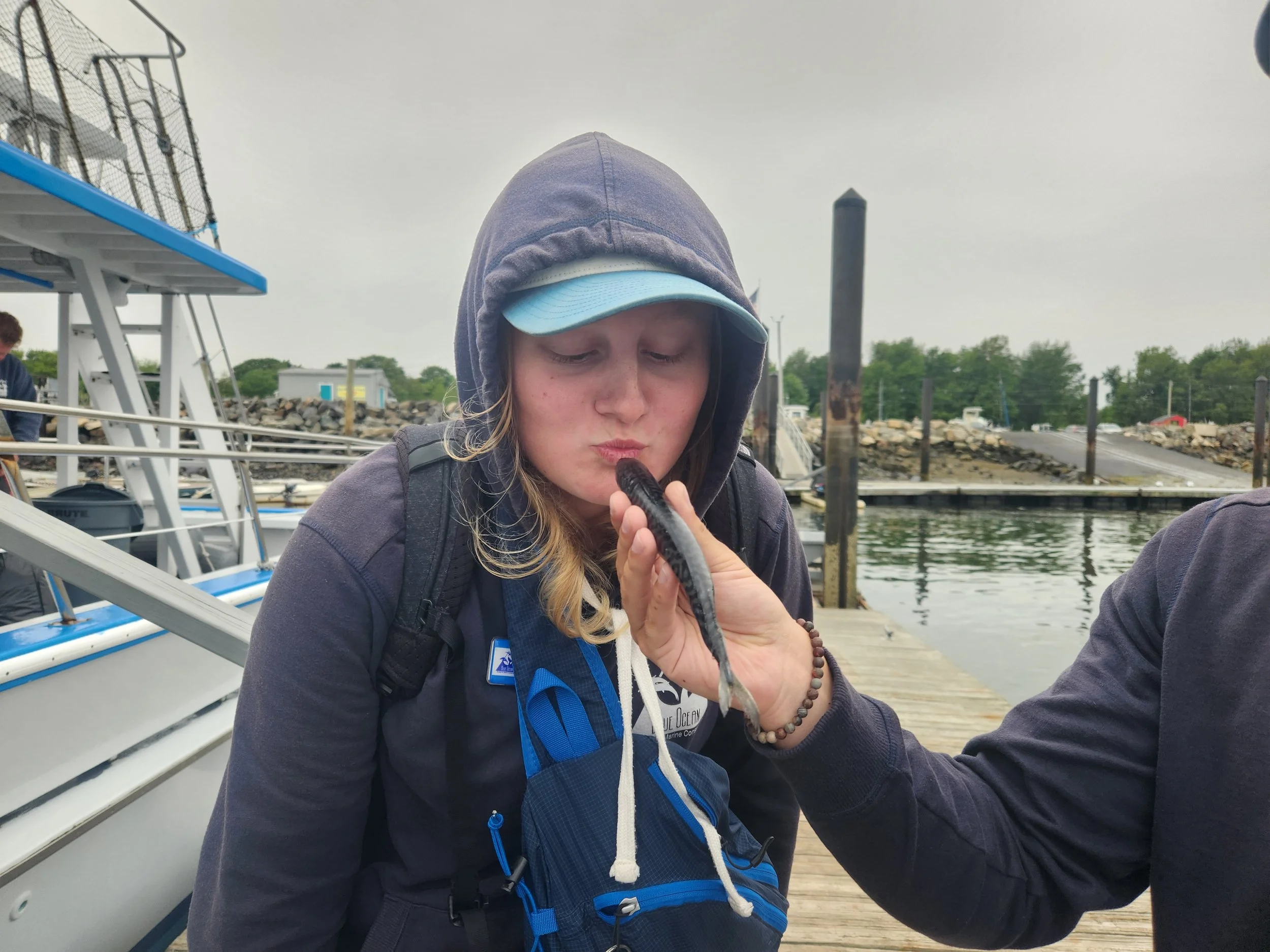 A young woman with blonde hair wearing a hoodie and cap, sitting on a dock at a marina, blowing into a small fish, with boats, rocks, and trees in the background.