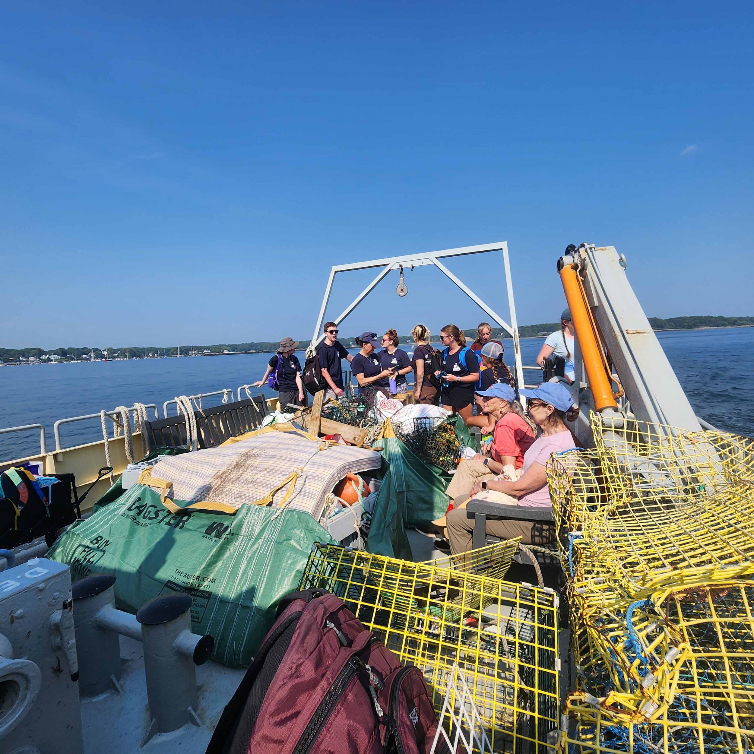 People on a boat filled with gear, backpacks, and supplies, with a clear blue sky and a waterfront in the background.