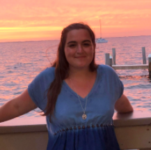 Girl smiling on boat at sunset with water and a sailboat in the background.