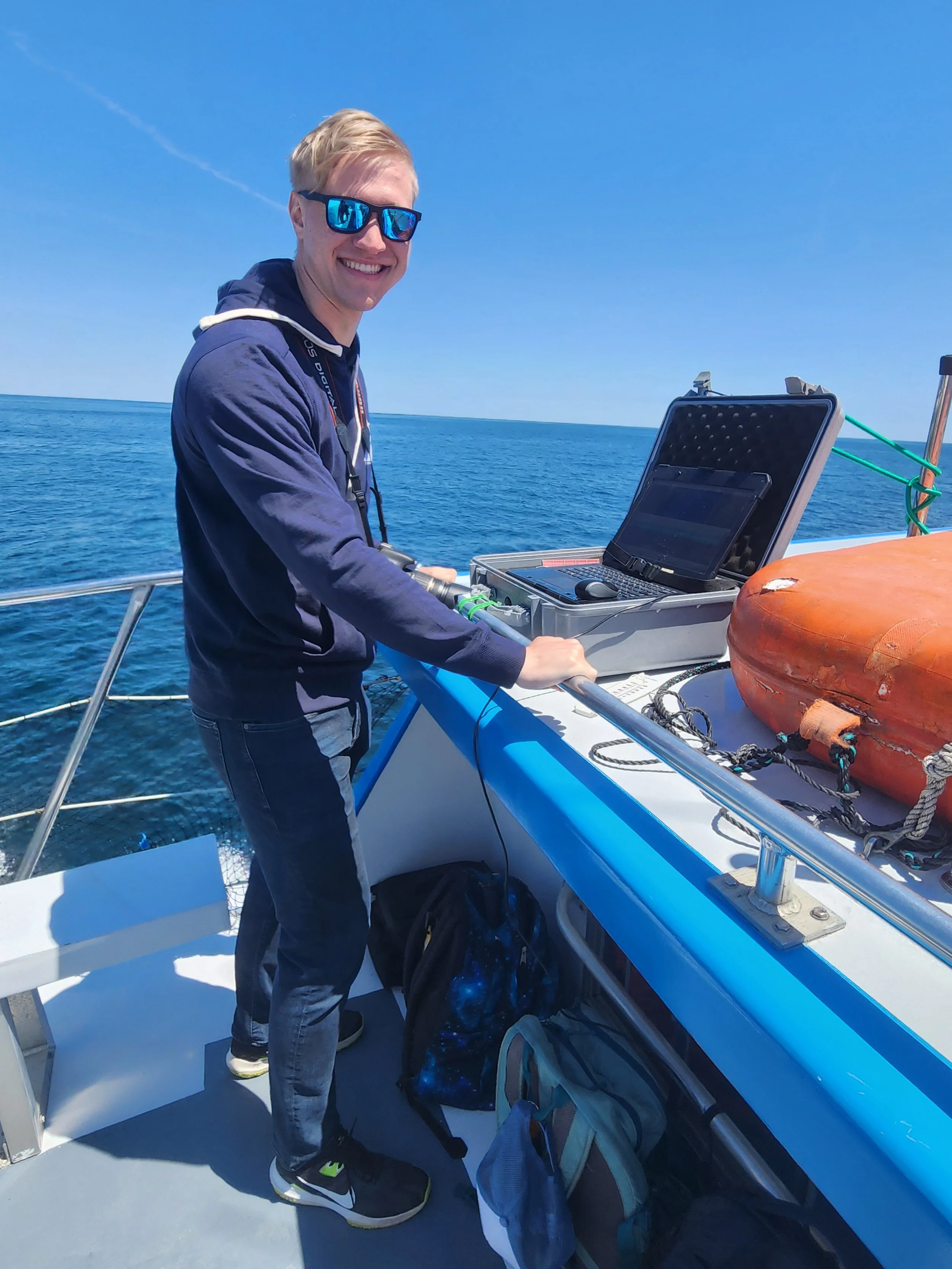 A young man with blond hair wearing sunglasses and a dark jacket is smiling on a boat with the ocean in the background. There is a laptop open on the boat's surface and some bags and equipment nearby.
