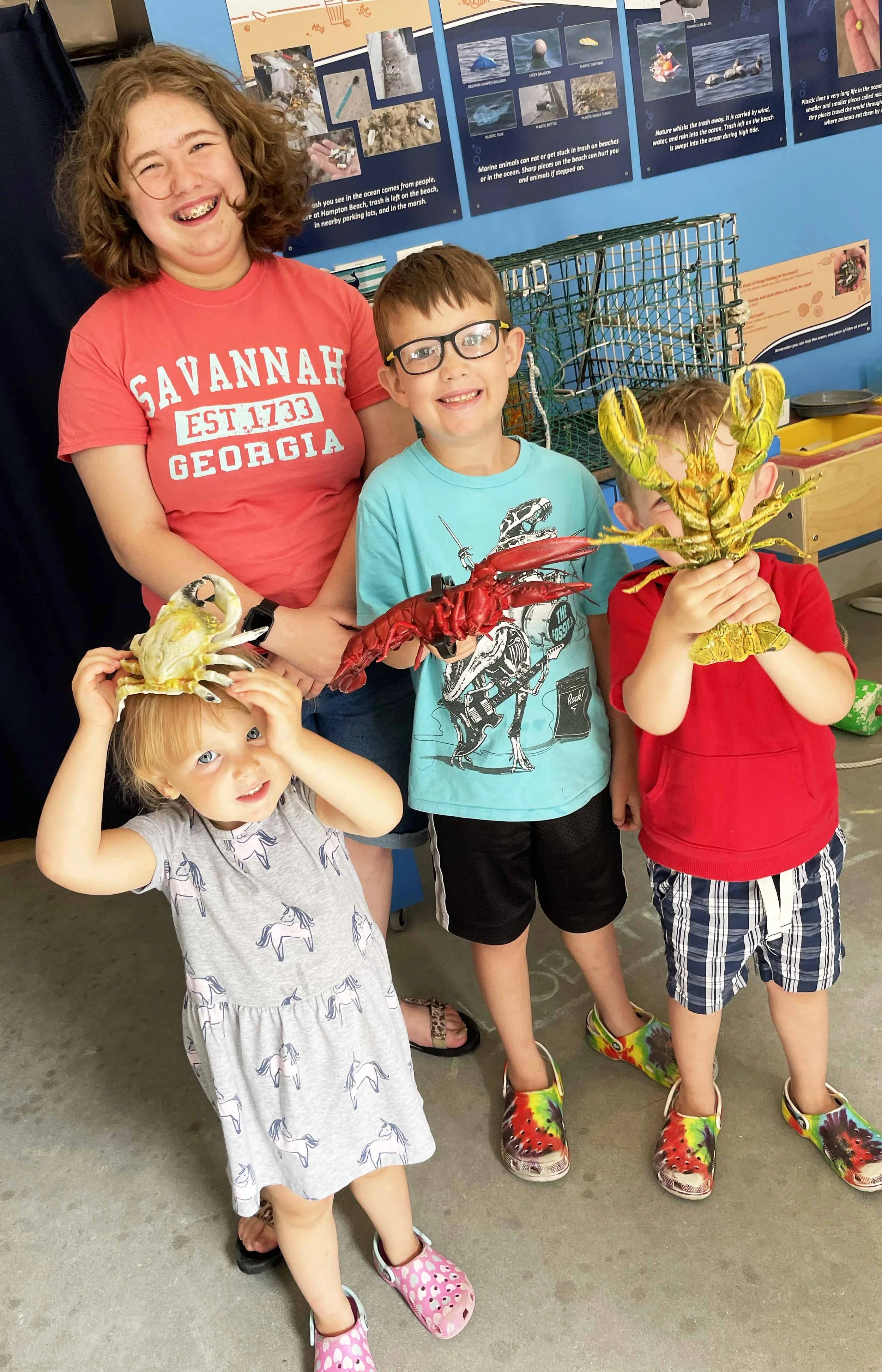 Four children are posing indoors, holding large toy lobsters and crabs. One girl is smiling widely, wearing a dress with horses on it and pink sandals. A boy with glasses and a blue T-shirt is smiling. Two other children are behind them, one holding a yellow crab toy and the other a red lobster toy, both covering their faces with the toys. The background features posters about marine animals and a wire animal trap.