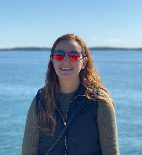 Woman with long hair wearing sunglasses and a vest, smiling outdoors near a body of water.