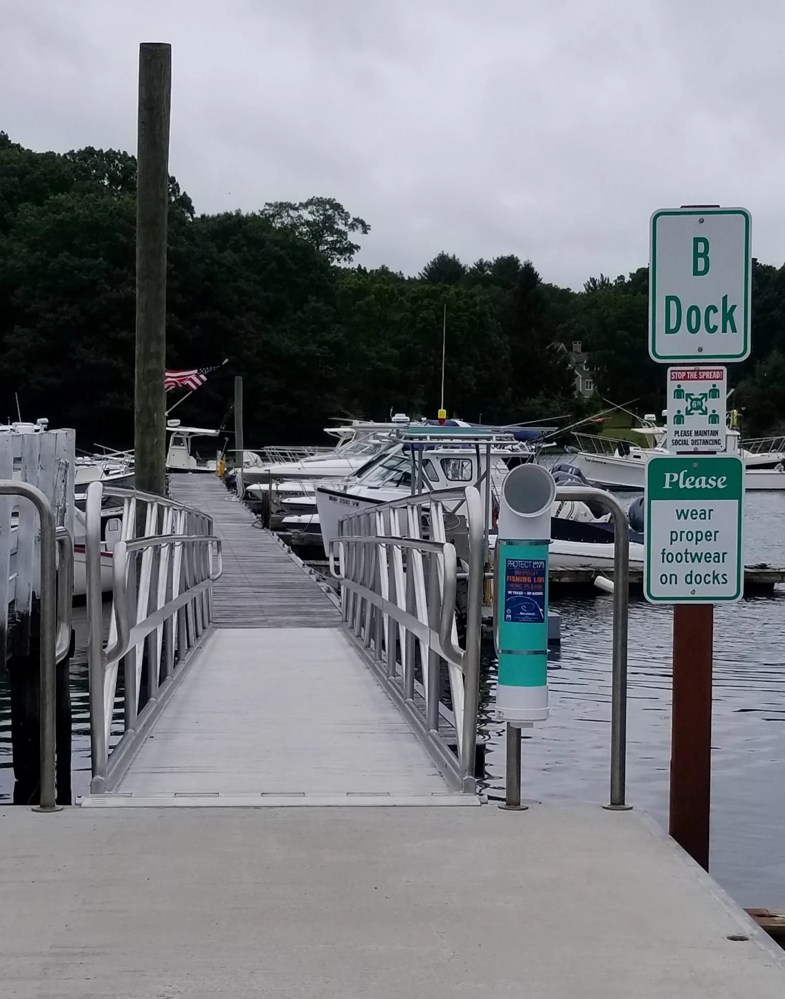 A marina with boats docked along the water, a wooden pier with railings leading to the boats, and a sign that says 'B Dock' with social distancing instructions. The sky is overcast and there are trees in the background.