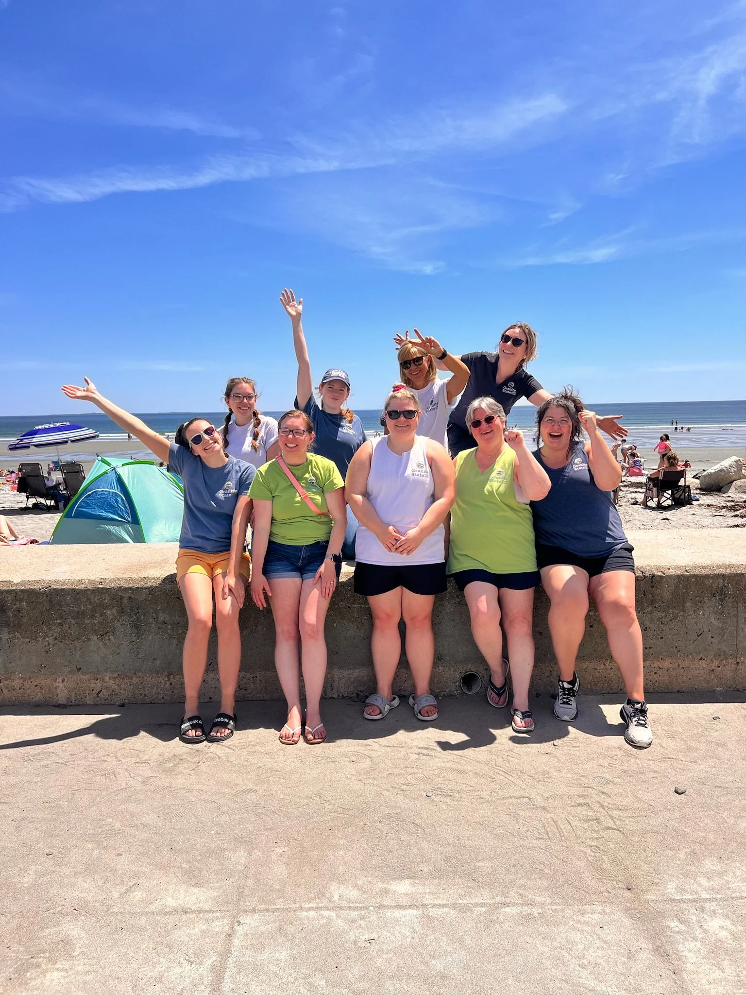 Group of nine women smiling and posing together on a beach, standing behind a concrete wall with ocean and blue sky in the background.