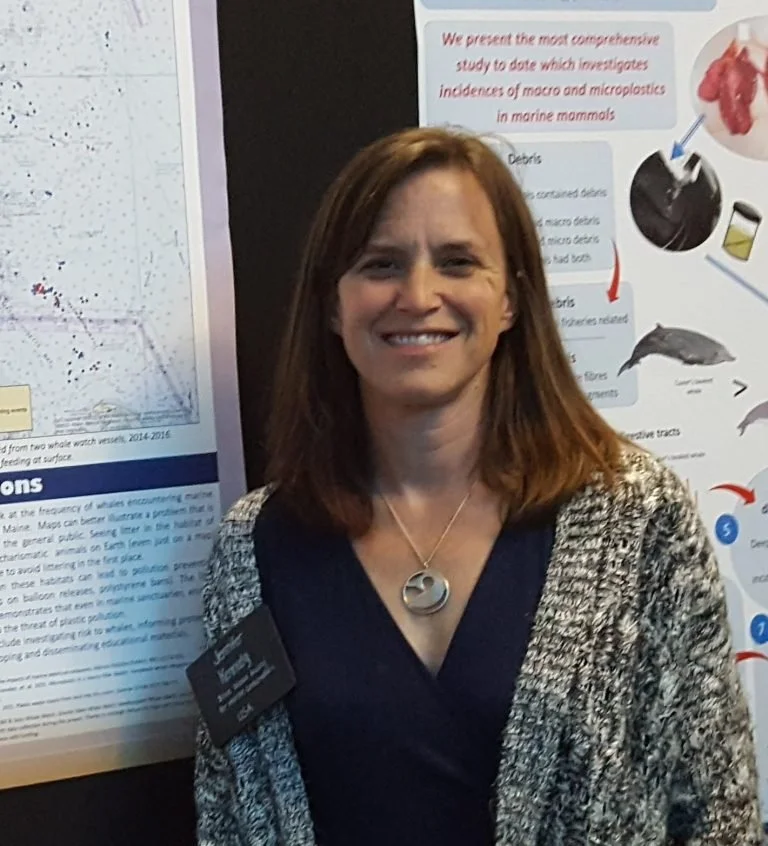 Woman smiling in front of scientific posters, wearing a black top, gray patterned cardigan, and a silver necklace with a circular pendant.