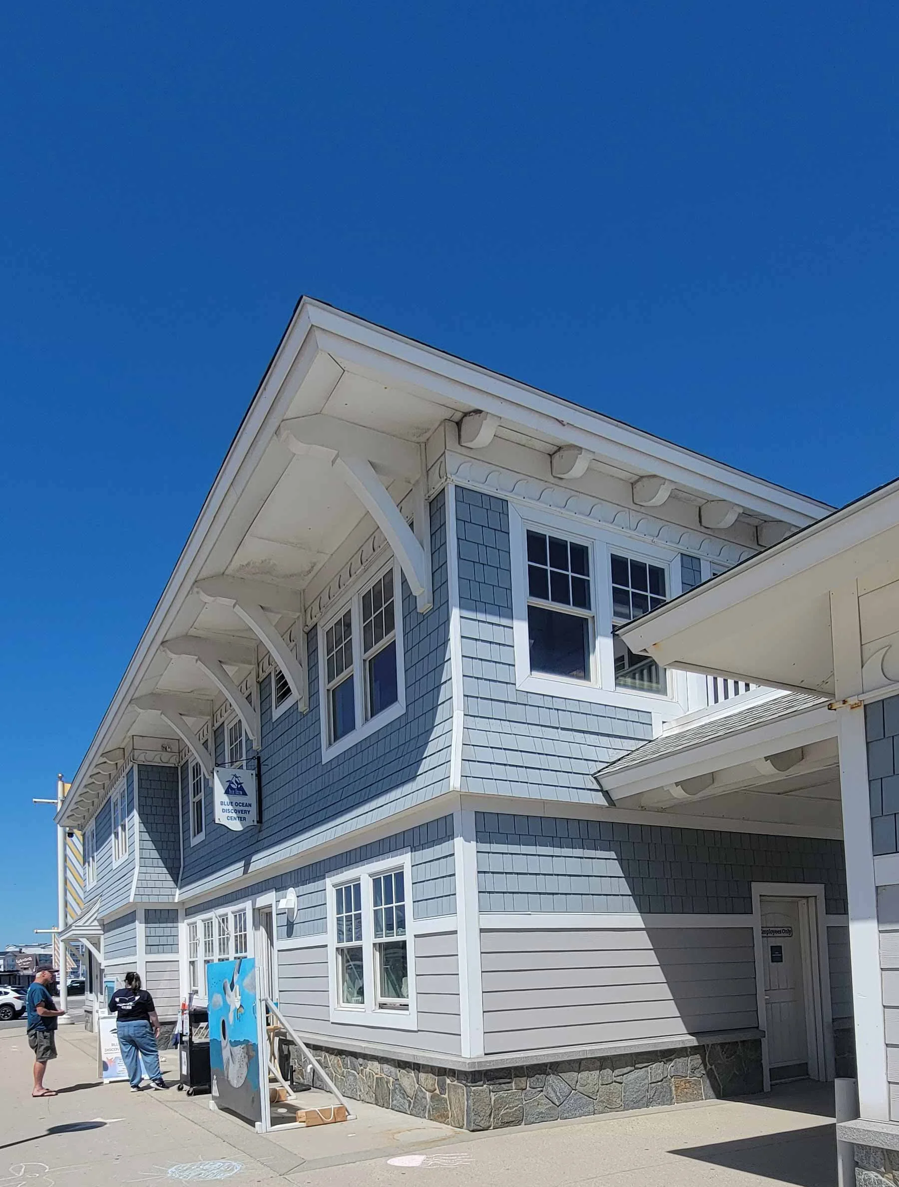 A two-story house painted in light blue with white trim and a stone foundation. The house has multiple windows, with some having small decorative overhangs, and features a sign that says "Blue Ocean Discovery Center" hanging from the upper part of the building. There are a few people standing outside under a bright blue sky.
