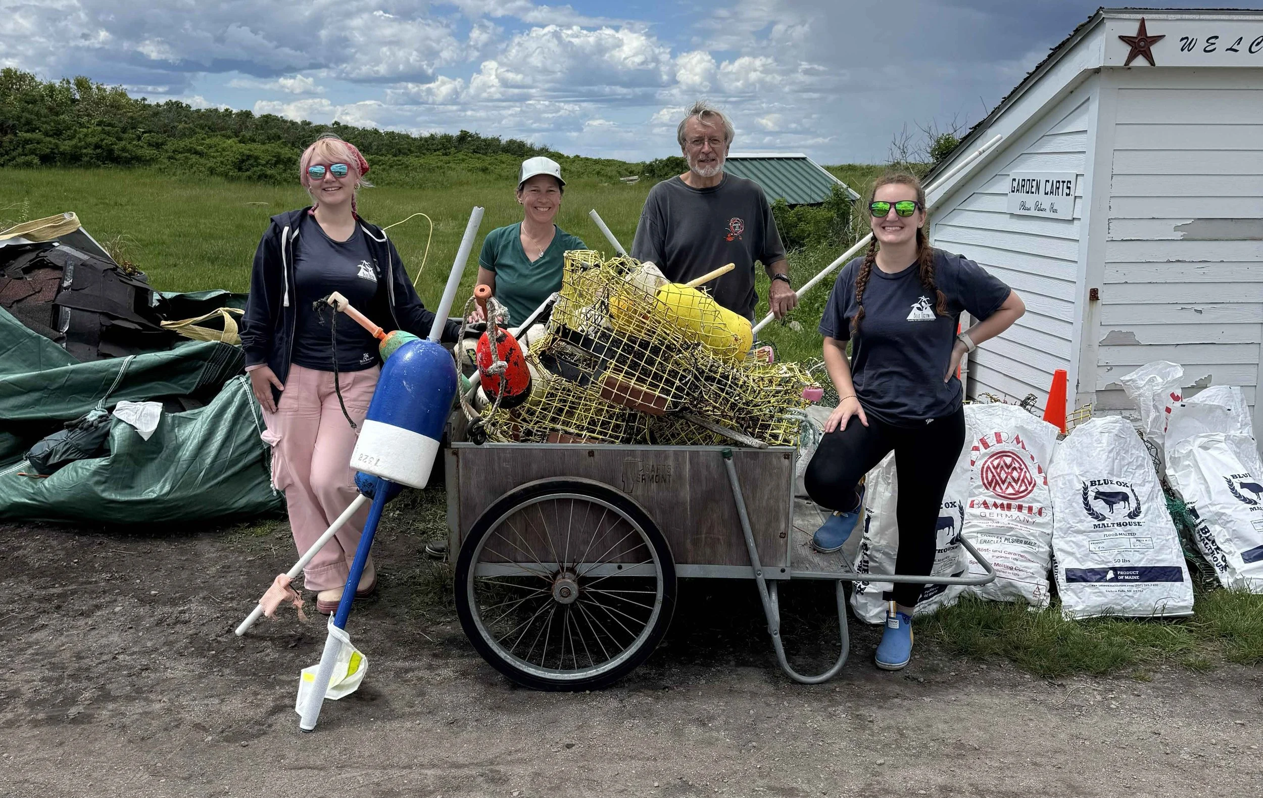 Four people standing outdoors near a cart filled with trash and recyclables. They are smiling, wearing casual clothes, and sunglasses. There is a small white building with a sign that reads 'Garden Carts' behind them, and piles of bags labeled with various recycling brands on the ground. The background includes green grass, trees, and a partly cloudy sky.