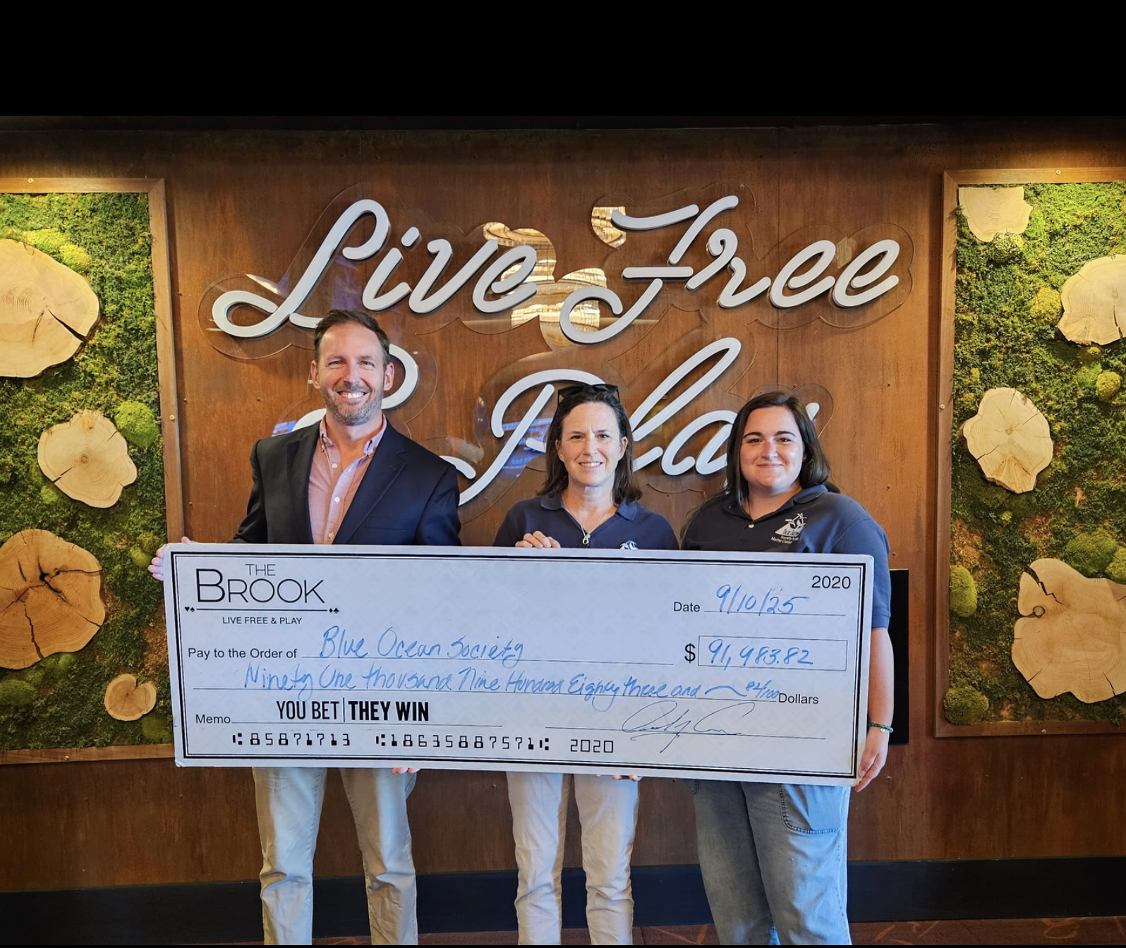 Three people standing together holding a large check in front of a wall with a neon sign that reads 'Live Free, Ride Free'.