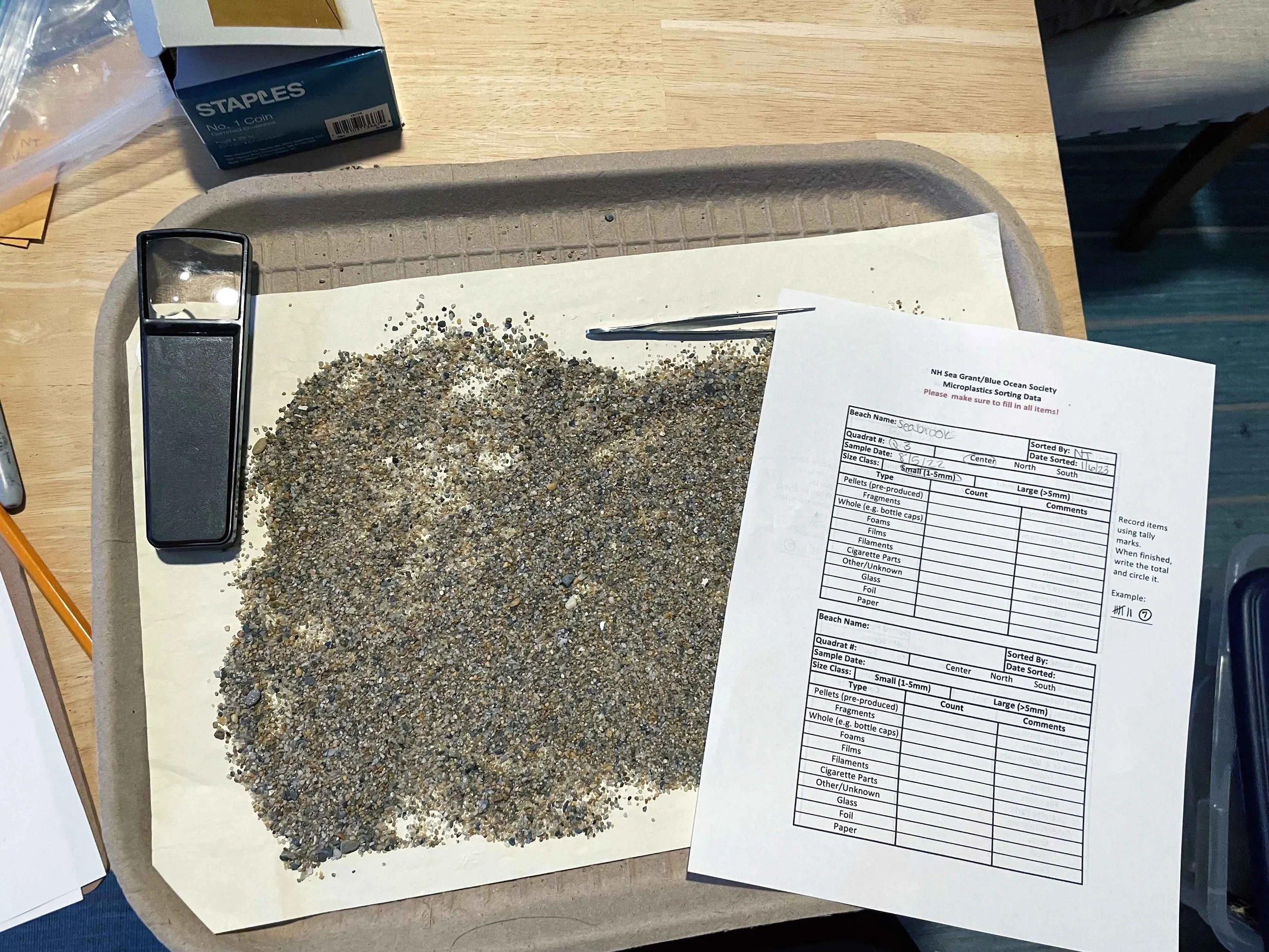 A tray with a pile of small rocks or gravel and a pair of tweezers, with a sheet of paper containing data and a magnifying glass nearby, on a wood table.