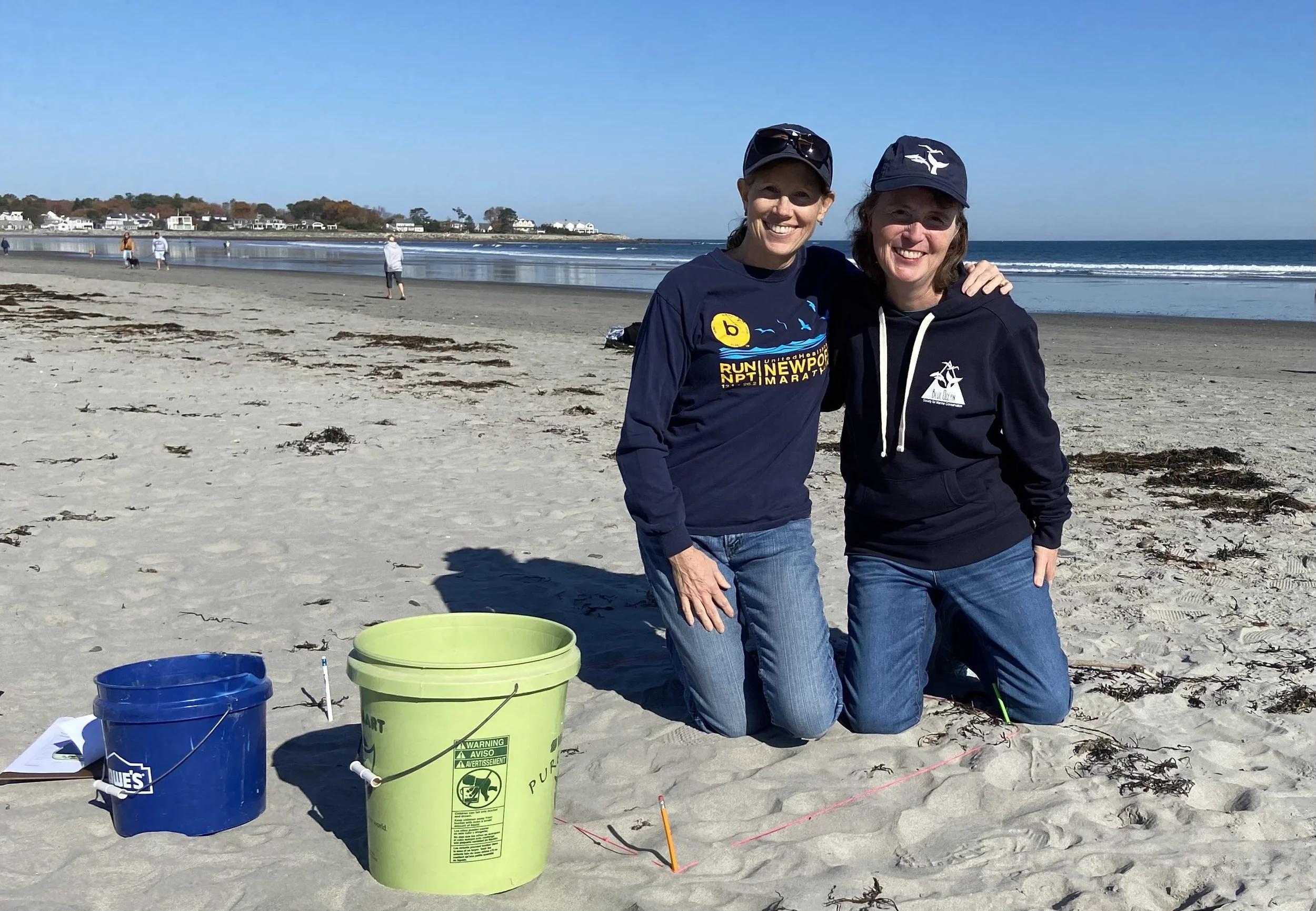 Two smiling women kneeling on a sandy beach with buckets, in front of dotted fishing lines, with the ocean and a clear sky in the background.