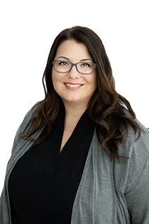 Portrait of a woman with dark hair wearing glasses, a black top, and a gray blazer, smiling against a white background.