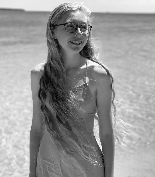 A woman with long wavy hair, wearing glasses, smiling on a beach with water in the background.