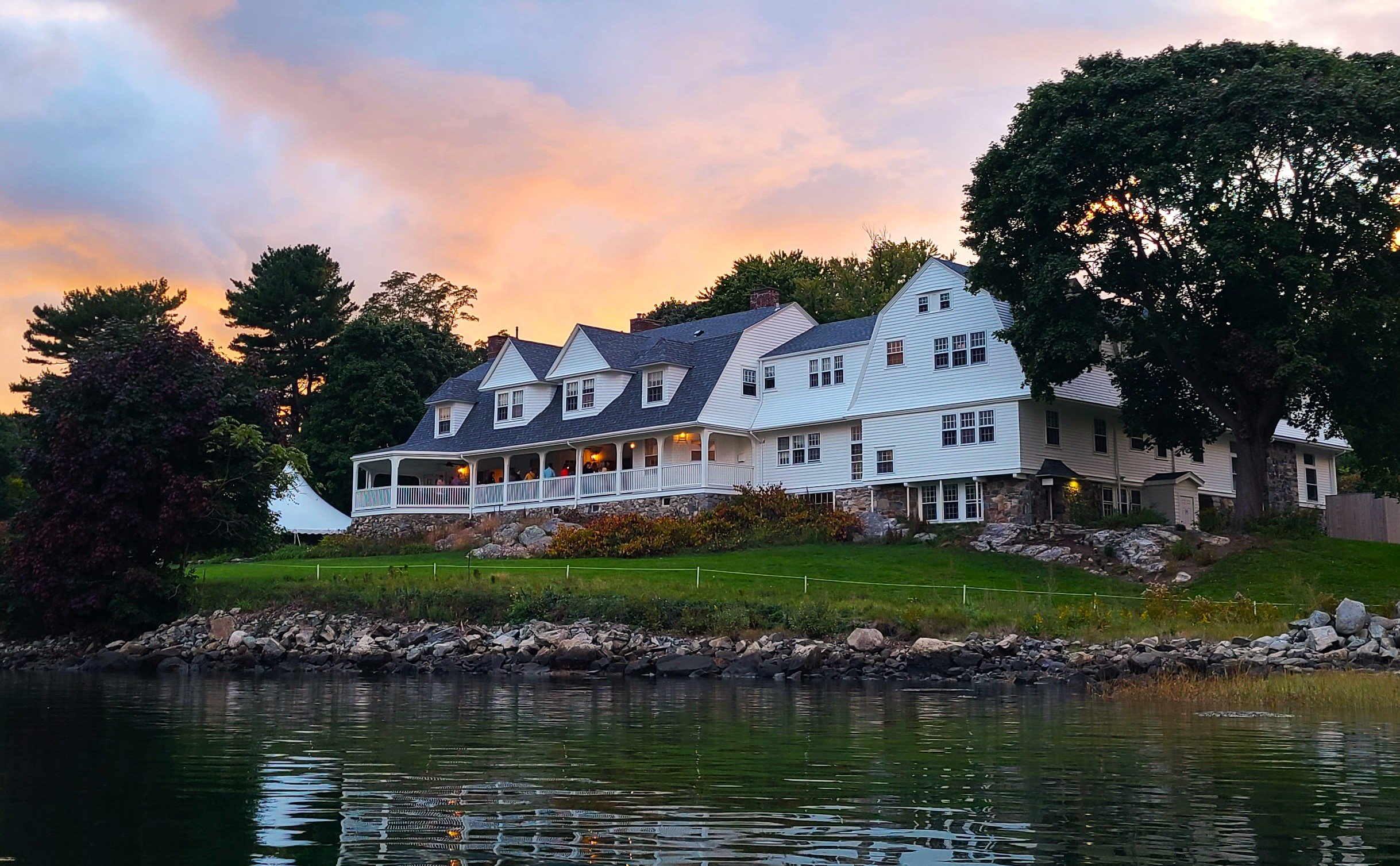 A large white house with water in the foreground and the sunset in the background