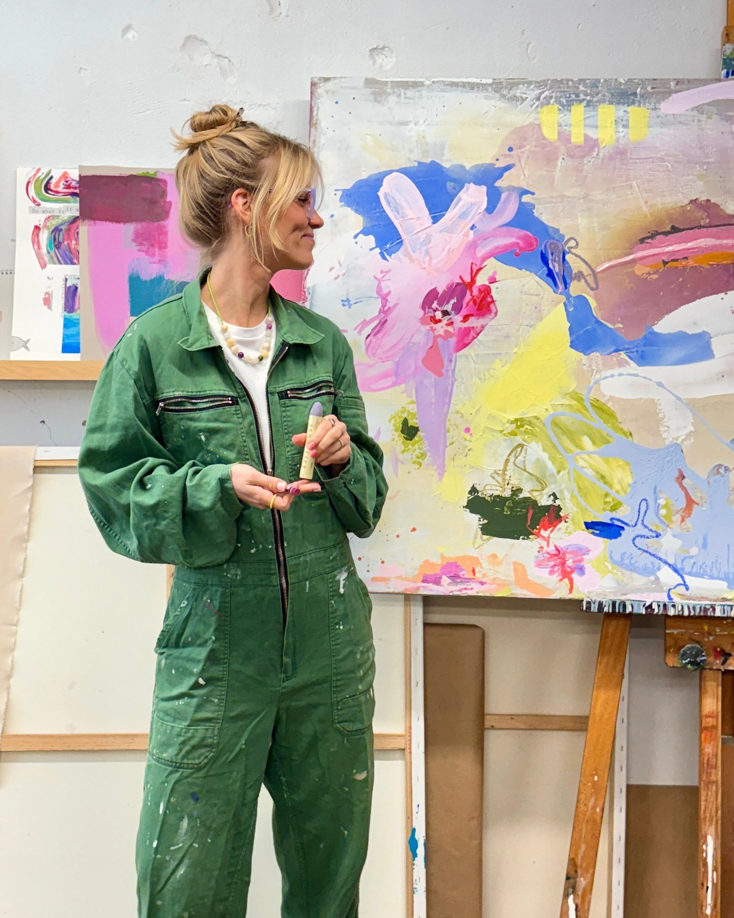 Eine Frau mit Brille und hellblondem Haar in einem Dutt, trägt eine grüne Maljacke mit einem weißen T-Shirt darunter, steht vor einer abstrakten bunten Leinwand in einem Kunststudio.