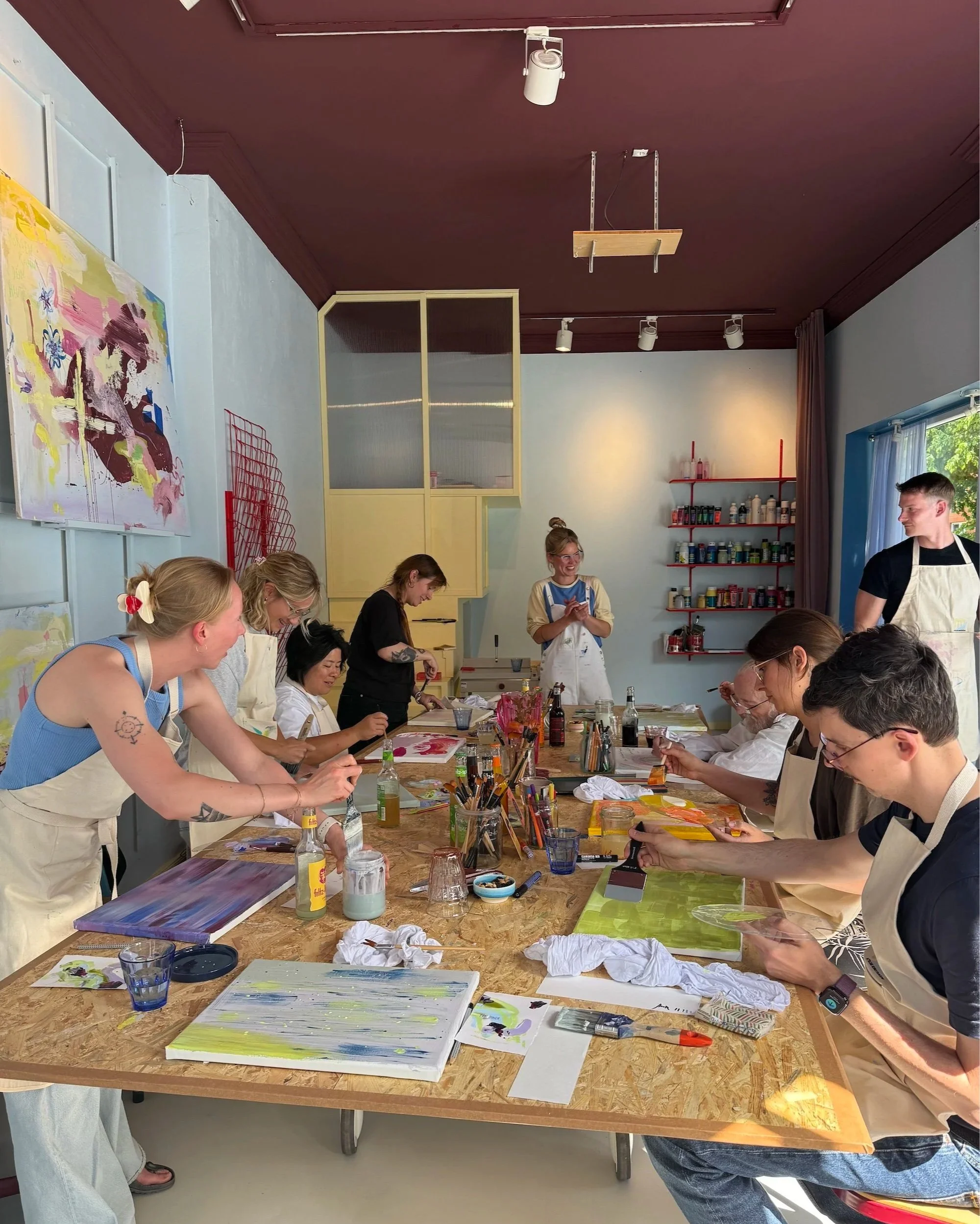 Menschen beim Malworkshop in einem Raum, mit bunten Bildern, Staffeleien, Pinseln und Farben auf einem Tisch