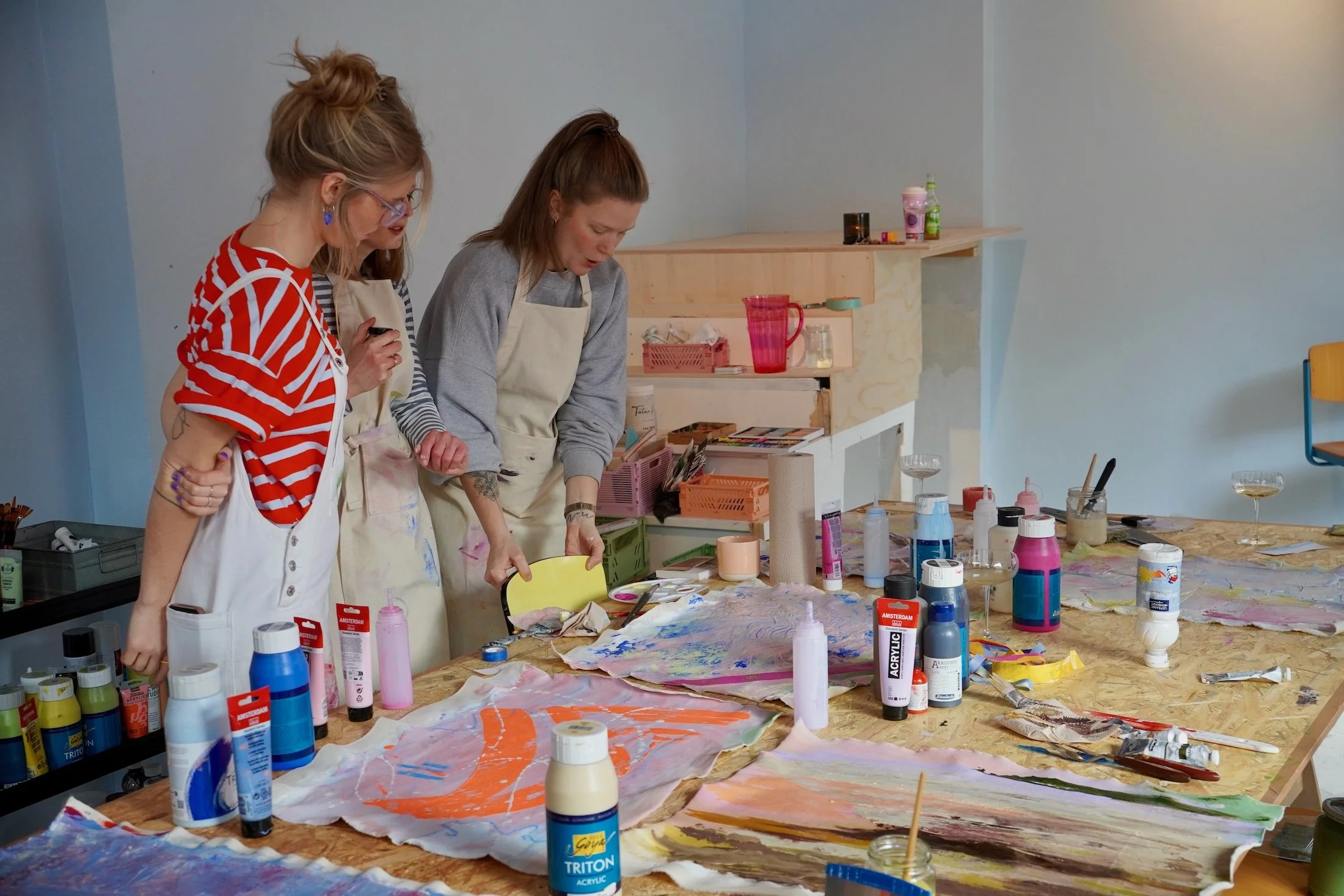 Drei Frauen beim Malen in einem Kunstatelier, umgeben von Farben, Pinseln und Kunstmaterialien.