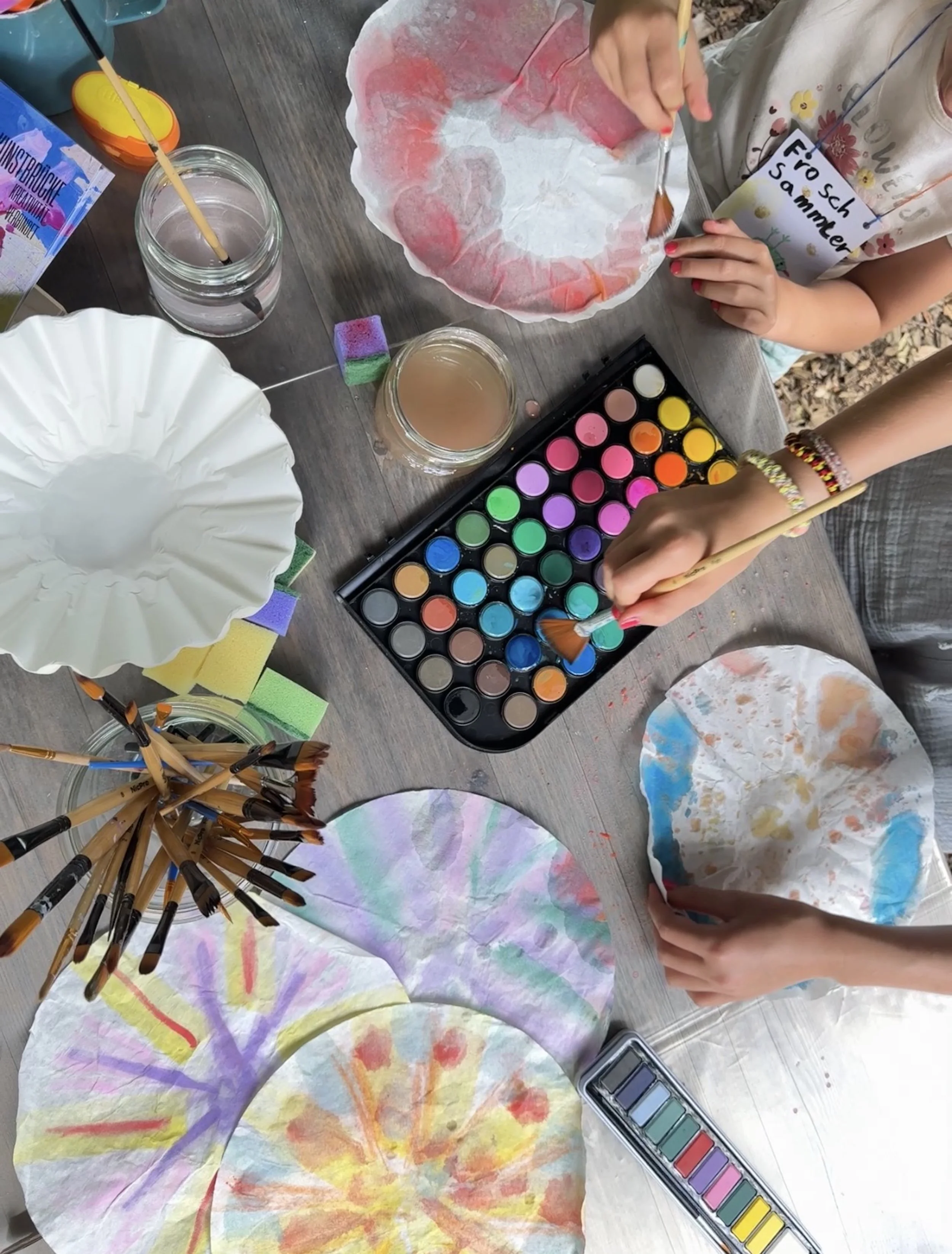 Kinder malen bunte Regenbögen auf Papier, mit Wasserfarben, Pinsel, Schwämmen, und Malmaterialien auf einem Tisch.