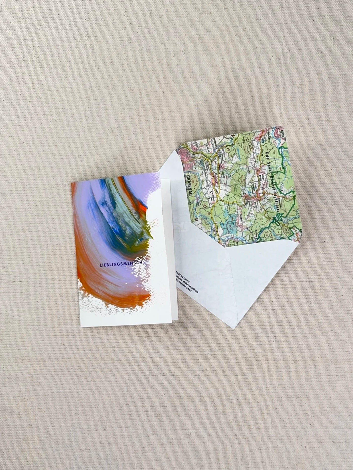 Zwei gefaltete Karten, eine mit einer topografischen Karte und eine mit einer Kunstzeichnung, liegen auf einem beige Stoffhintergrund.