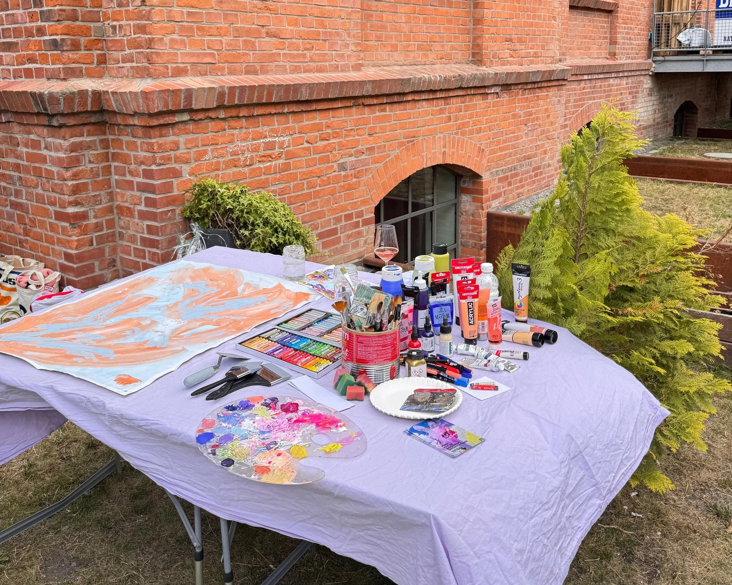 Ein Tisch im Freien mit Mal- und Zeichenmaterialien, darunter Farben, Pinsel, Leinwände und eine Palette, vor einer roten Backsteinwand.