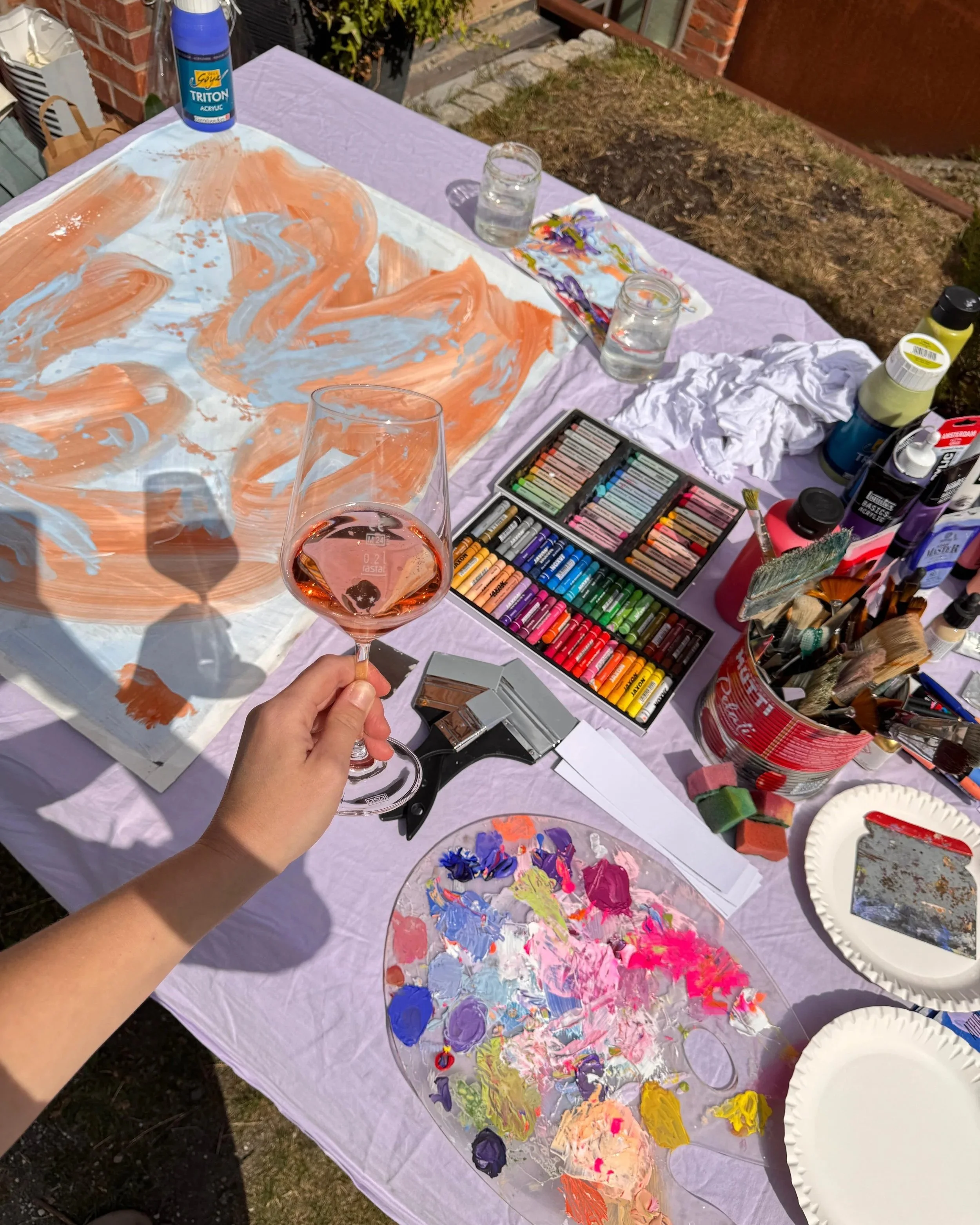 Ein Tisch mit Malutensilien, einer Palette mit bunten Farben, einem Glas mit Wasser, färbige Papierbappen, verschiedenen Acrylfarben und Pinsel, vermutlich für einen Malkurs im Freien.