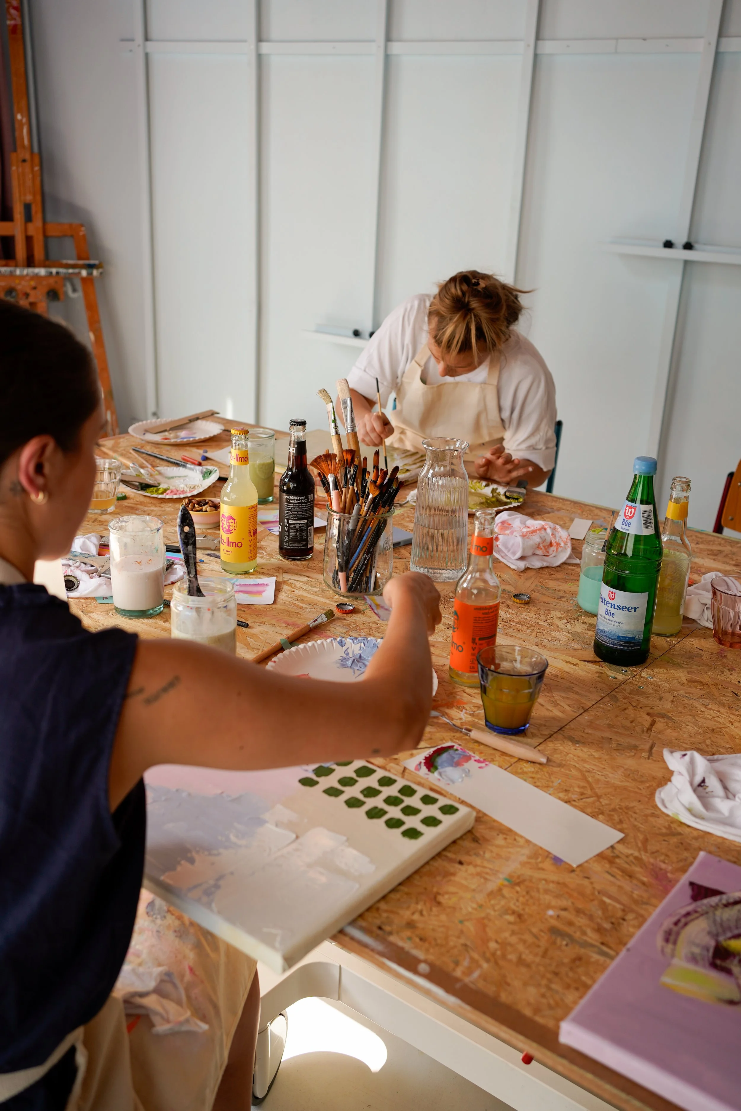 Menschen beim Malen, Sitzende Person in Vordergrund, Perlen und Kunstmaterialien auf dem Tisch, Künstler beim Arbeiten mit Farbpaletten und Pinseln, kreativ im Atelier.