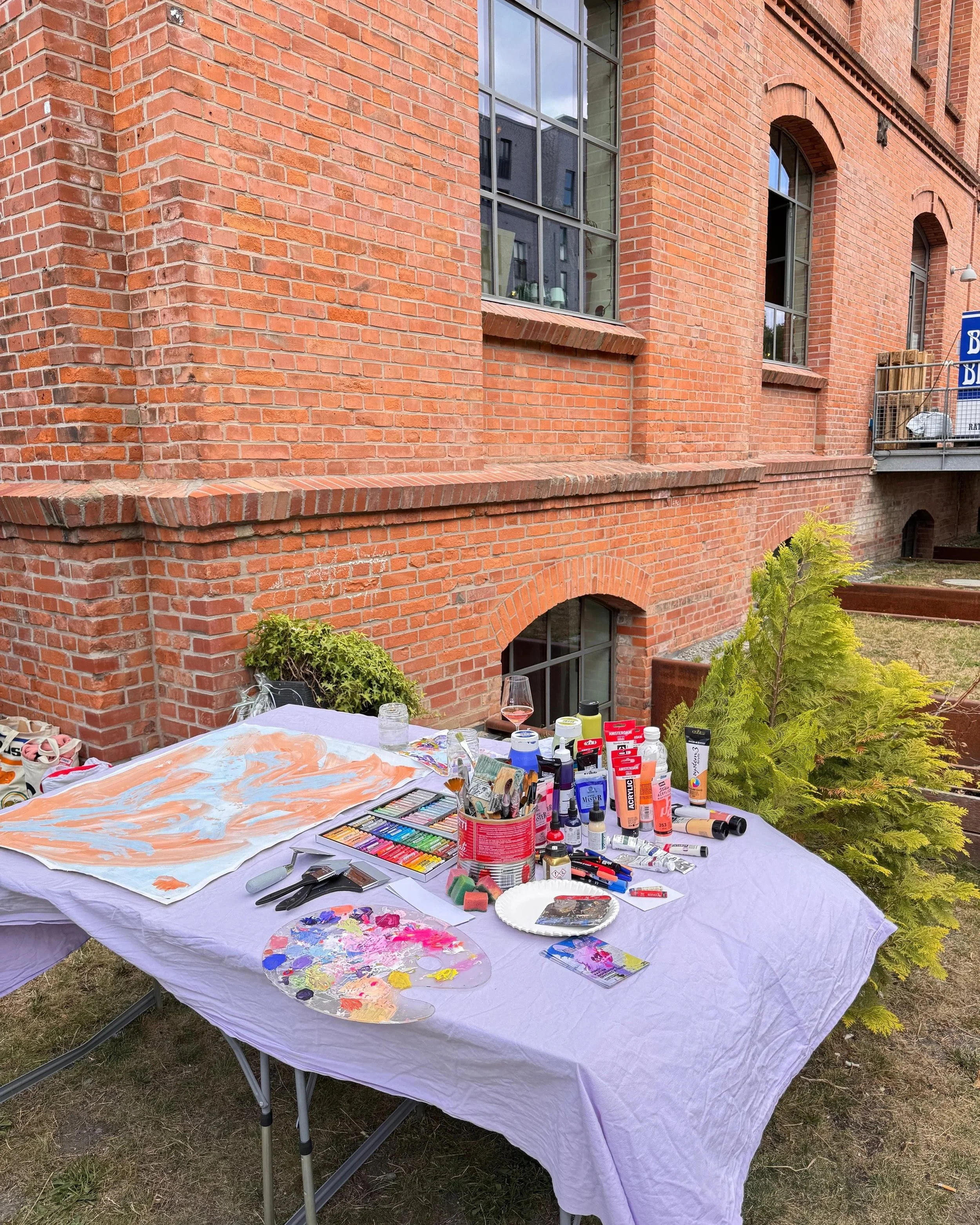 Ein Tisch im Freien mit Mal- und Bastelmaterialien, darunter Farbe, Pinsel, Skizzenbuch und andere Bastelutensilien, vor einer roten Backsteinwand.