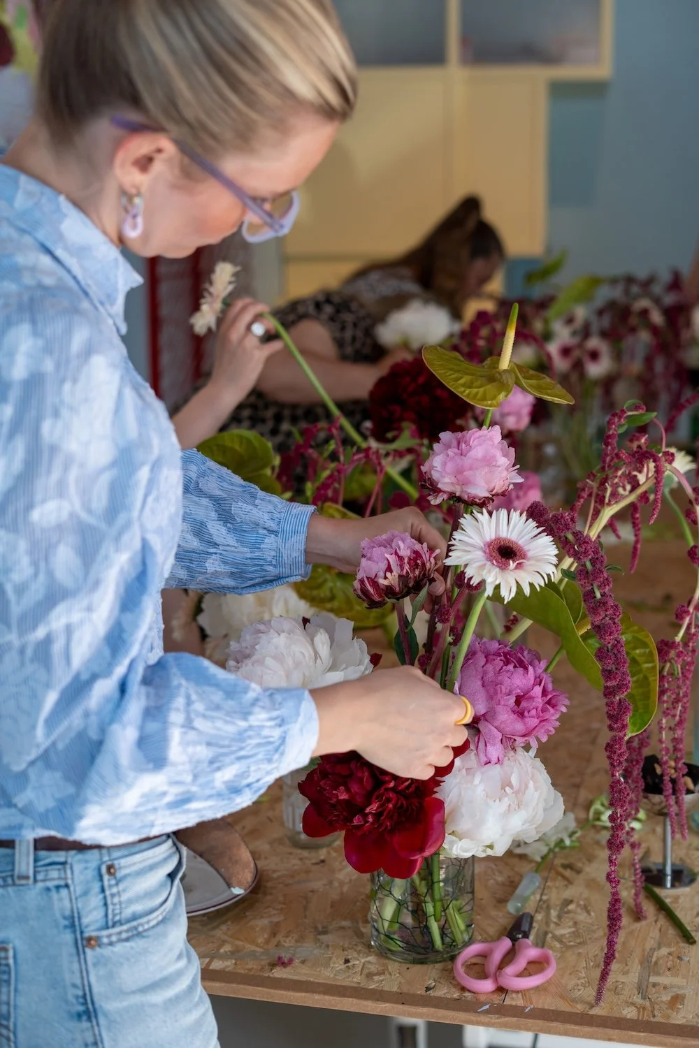 Frau arrangiert bunte Blumen in einer Vase auf einem Tisch, im Hintergrund weitere Blumen und eine zweite Frau.