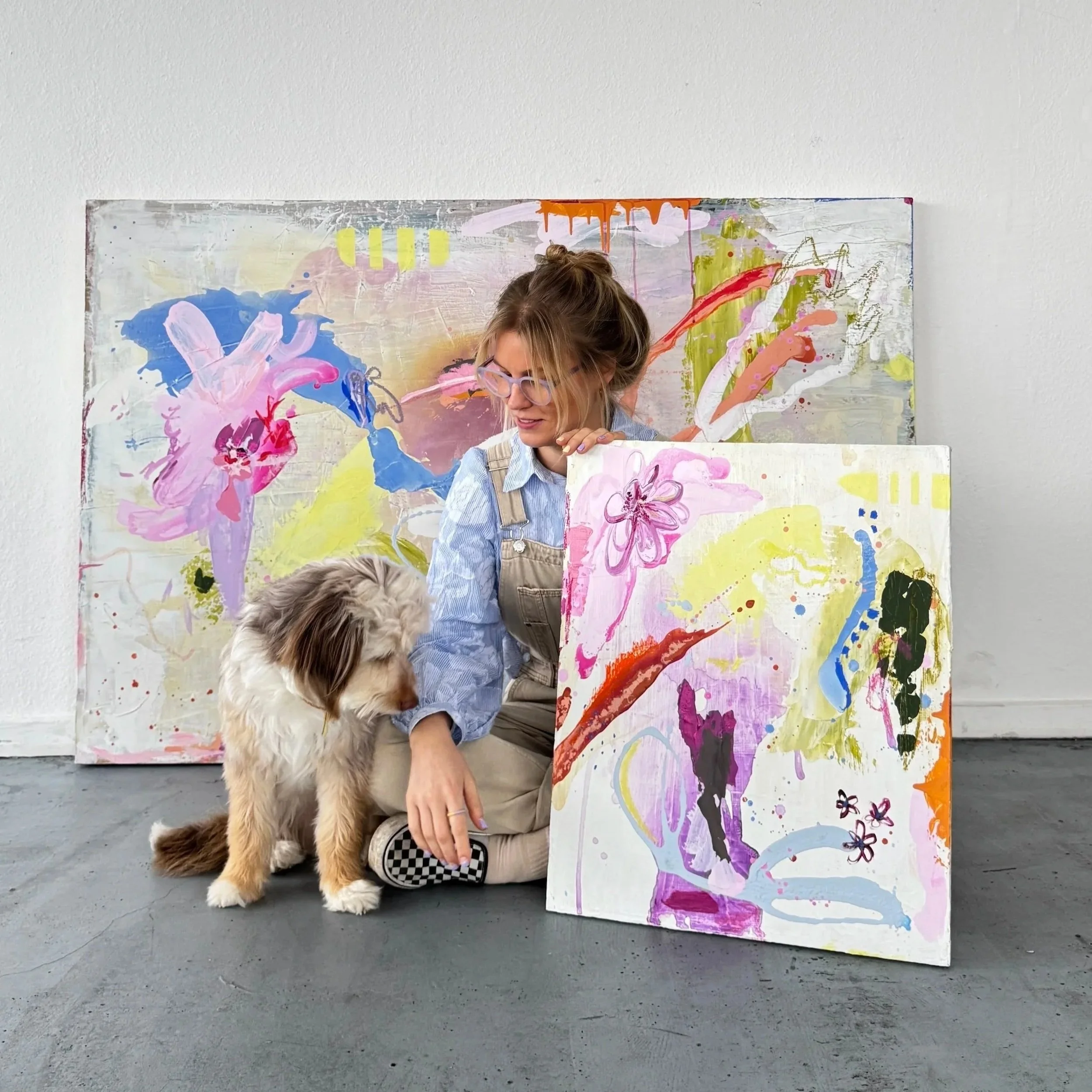 Ein Mensch sitzt auf dem Boden, hält eine große, bunte Leinwand, auf der Blumen und abstrakte Muster gemalt sind, und streichelt einen Hund. Im Hintergrund eine weitere große, bemalte Leinwand.