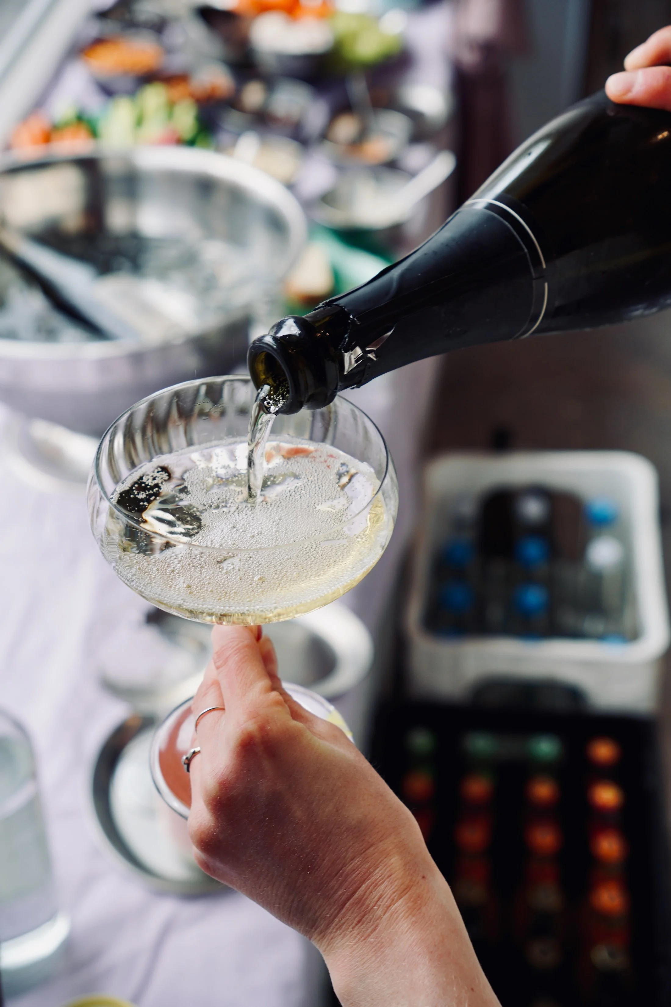 Eine Person gießt Sekt in ein Champagnerglas bei einer Feier oder einem festlichen Anlass, mit weiteren Gläsern und Essensständen im Hintergrund.