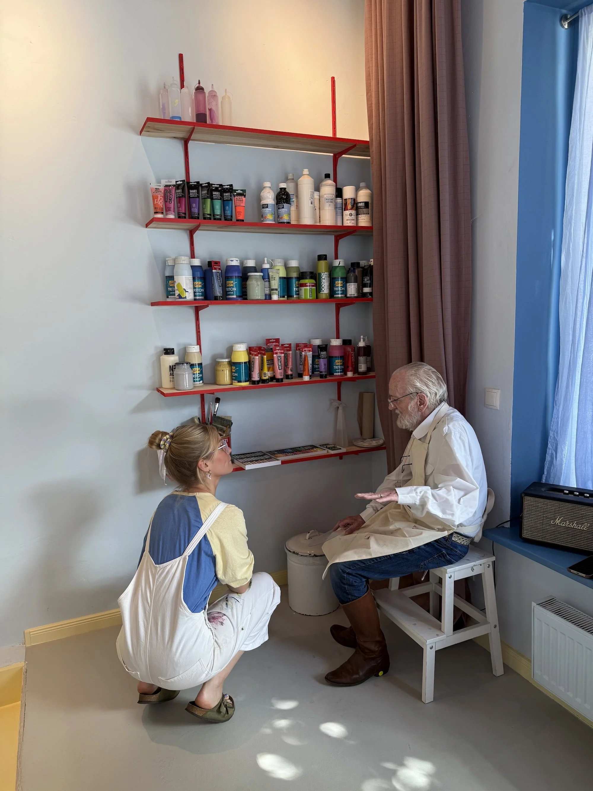 Ein älterer Mann sitzt auf einem Stuhl und spricht mit einem jüngeren, sitzenden Frauenpaar in einem Raum mit roten Wandregalen voller Tätowierfarben und -utensilien.