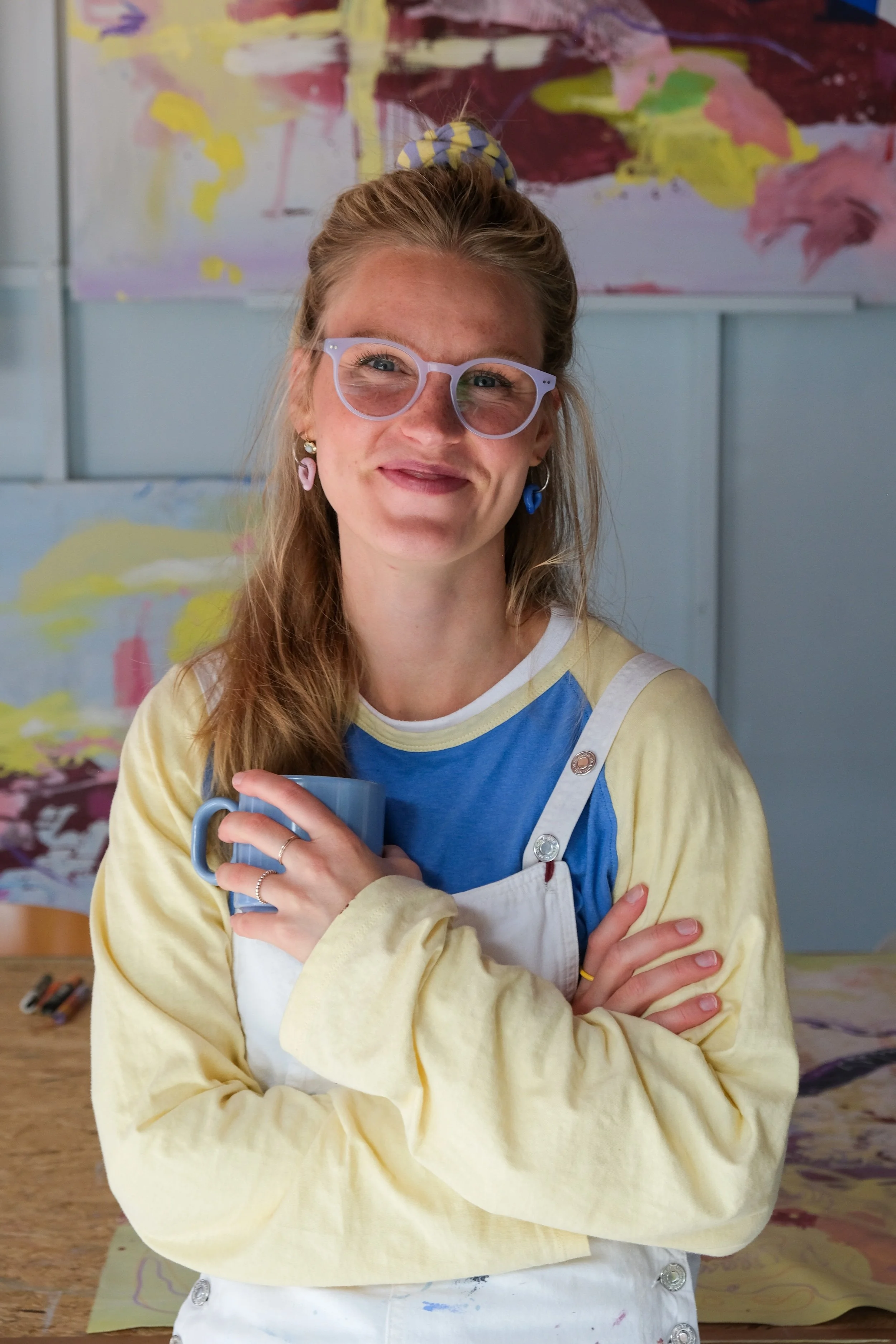 Eine lachende junge Frau mit roten Haaren, die eine Brille trägt, hält eine blaue Tasse in den Händen. Sie steht vor einem bunten, abstrakten Hintergrund in einem Raum, vermutlich während einer Kunstaktivität.