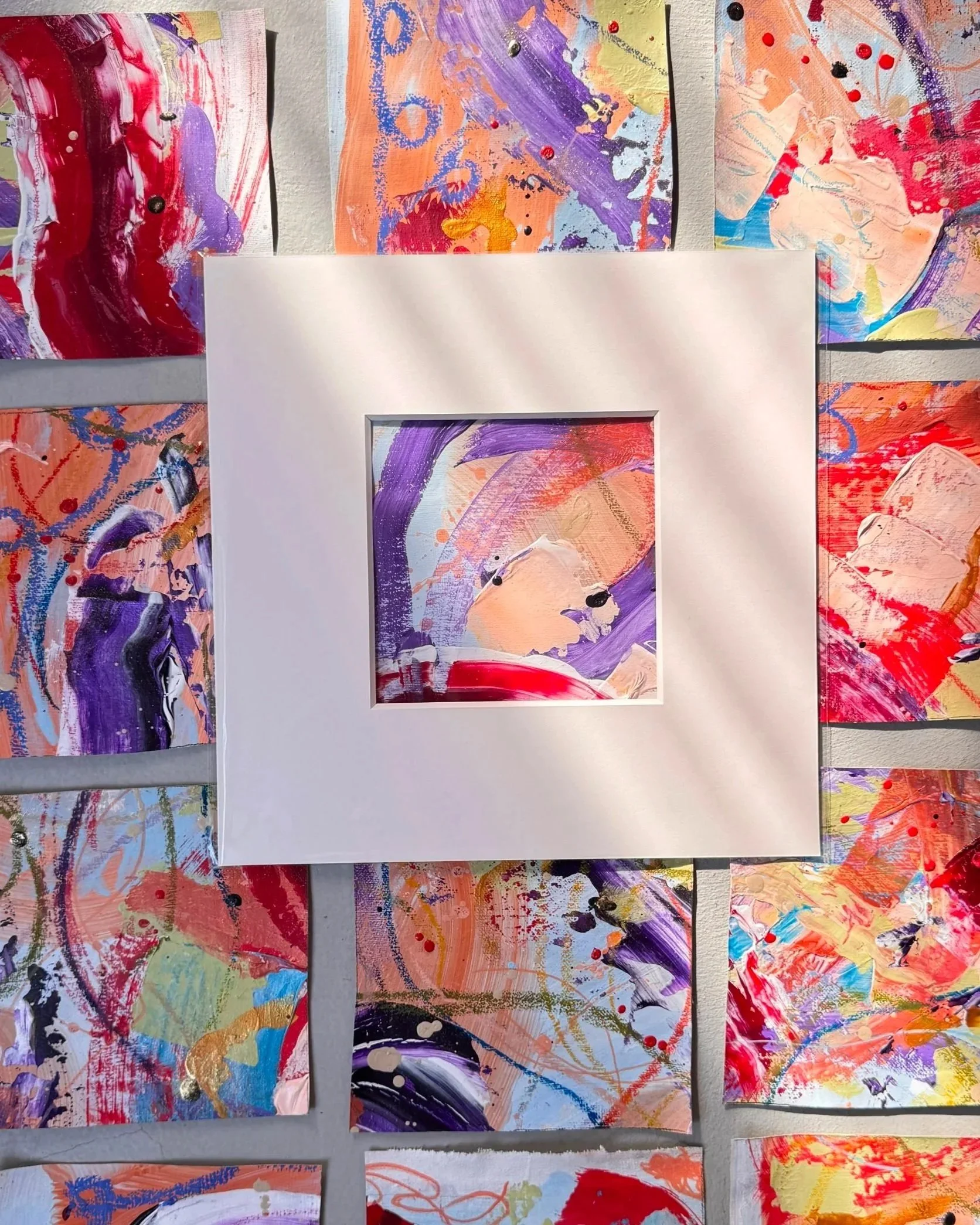 Abstrakte Kunstwerke, größtenteils in Rosa-, Lila- und Orangetönen mit Akzenten in Blau, Schwarz, Gold und Rot, sind auf einer Wand arrangiert. Die Gemälde bestehen aus dynamischen Pinselstrichen, Spritzern und Farbklecksen, wobei einzelne Werke teilweise durch weiße oder goldene Farben hervorgehoben werden.