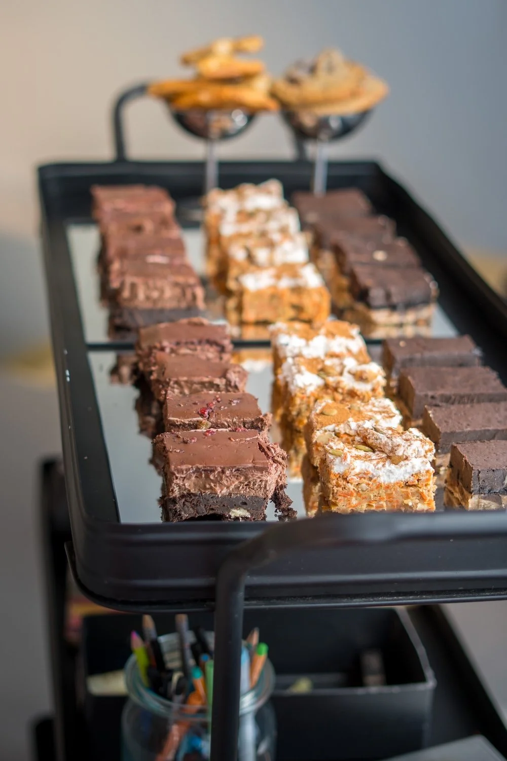 Verschiedene Sorten Kuchen auf einem schwarzen Servierwagen, im Hintergrund zwei glasierte Löffel mit Keksen.