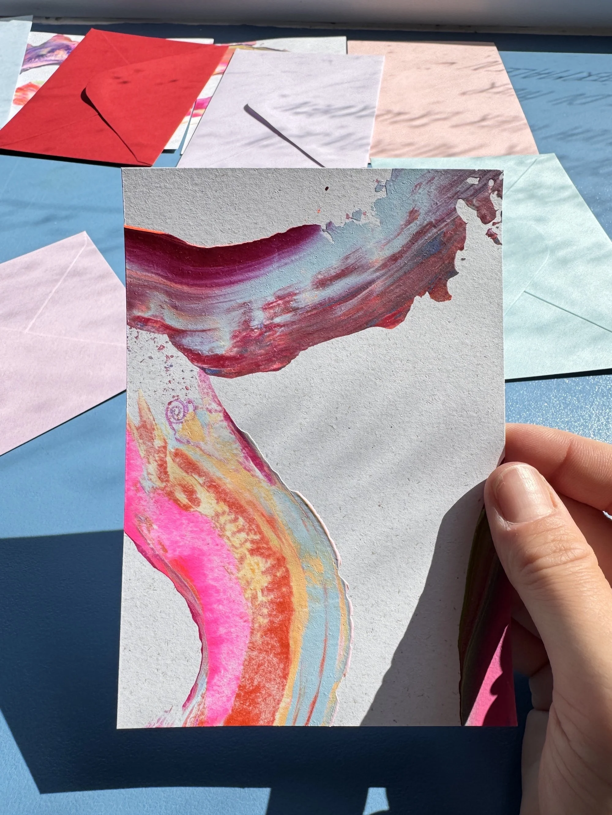 Bunte, abstrakte Aquarellmalerei auf einem Stück Papier, gehalten von einer Hand, mit weiteren Farbpaasten im Hintergrund, auf blauem Untergrund.