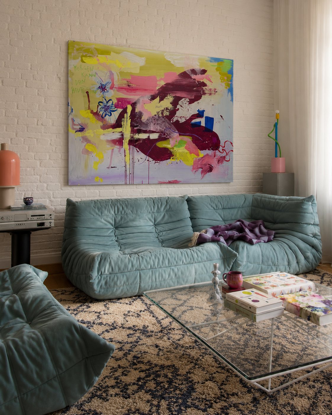 Moderner Wohnzimmerbereich mit Blumenmuster-Sofa, abstrakter bunter Wandkunst, Couchtisch mit Büchern, Kerzen, und Dekorationsgegenständen, Teppich, seitlicher Lampenständer und Vorhänge an Fenster mit natürlichem Licht.