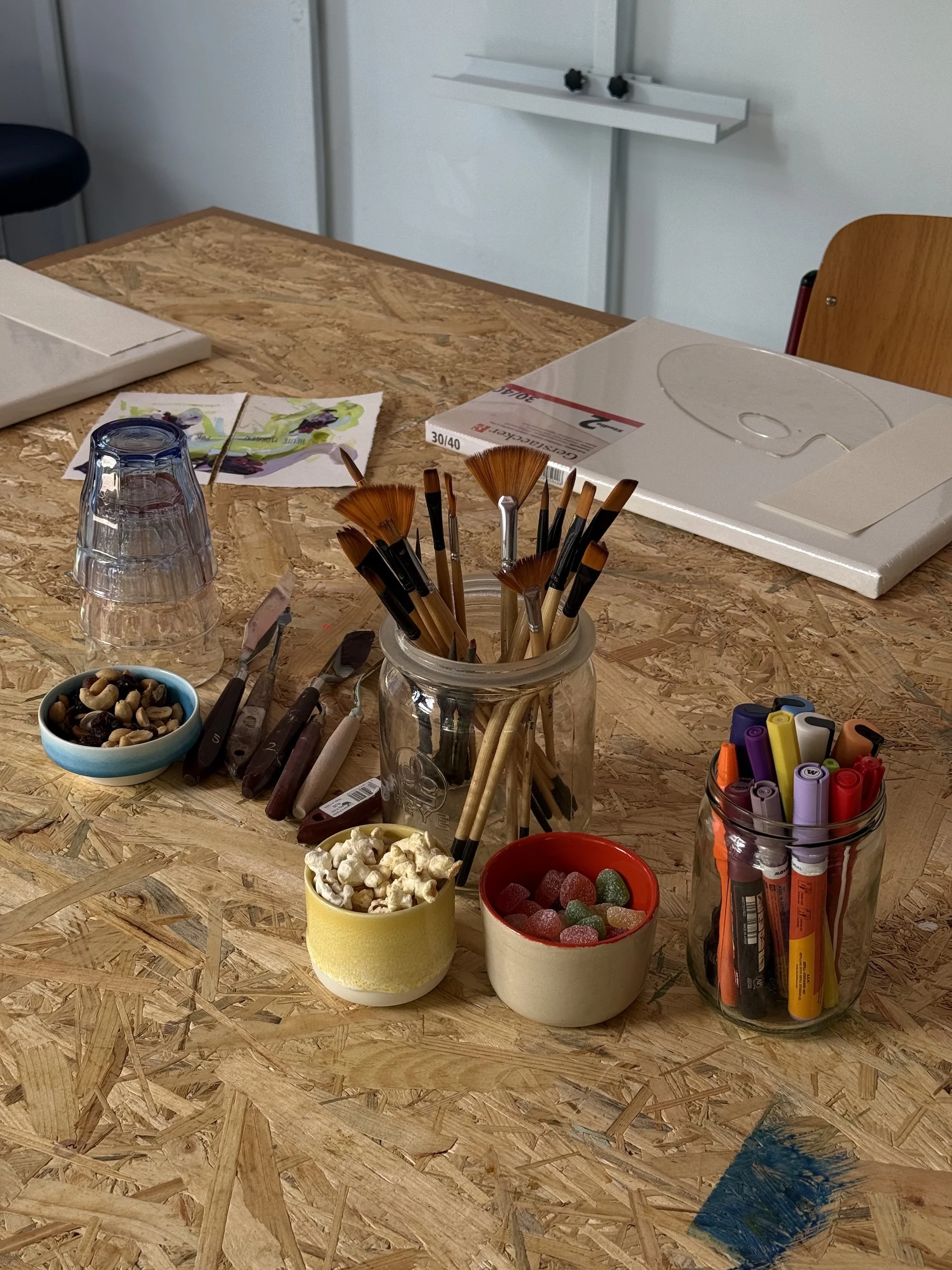 Ein Tisch mit verschiedenen Mal- und Bastelmaterialien, darunter Pinsel, Marker, Süßigkeiten, eine leere Trinkschale und ein Pappkarton.