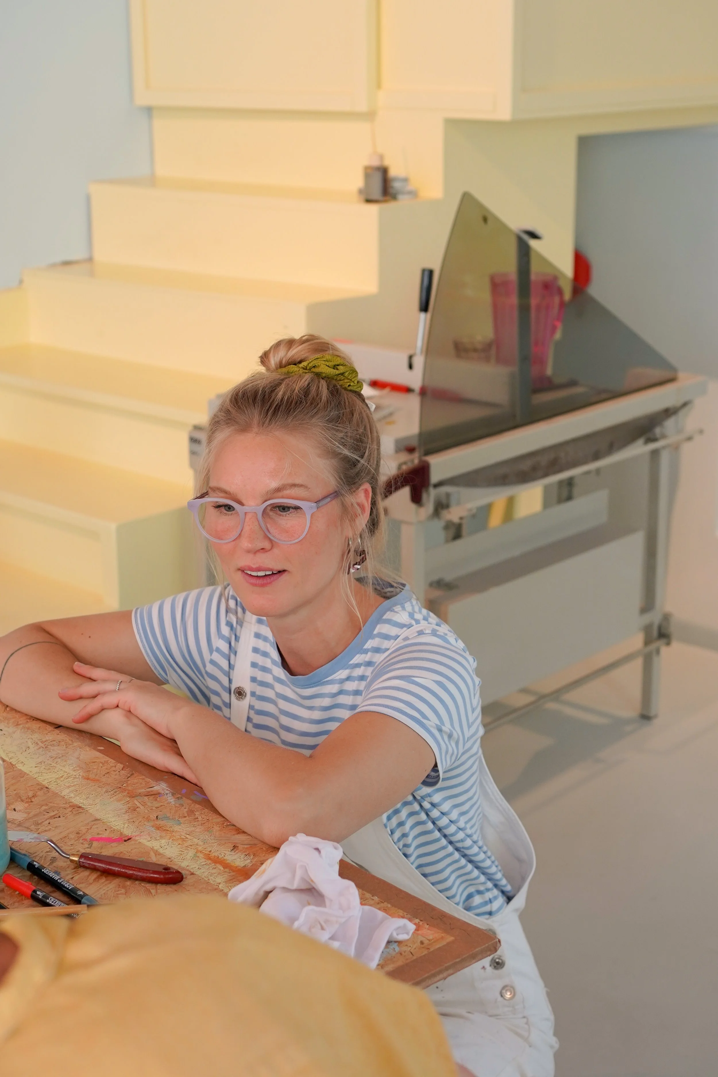 Junge Frau mit Brille und Zopf, sitzt an einem Tisch im Werkraum, während sie an einem Projekt arbeitet, mit Werkzeugen und Stoffresten auf dem Tisch.