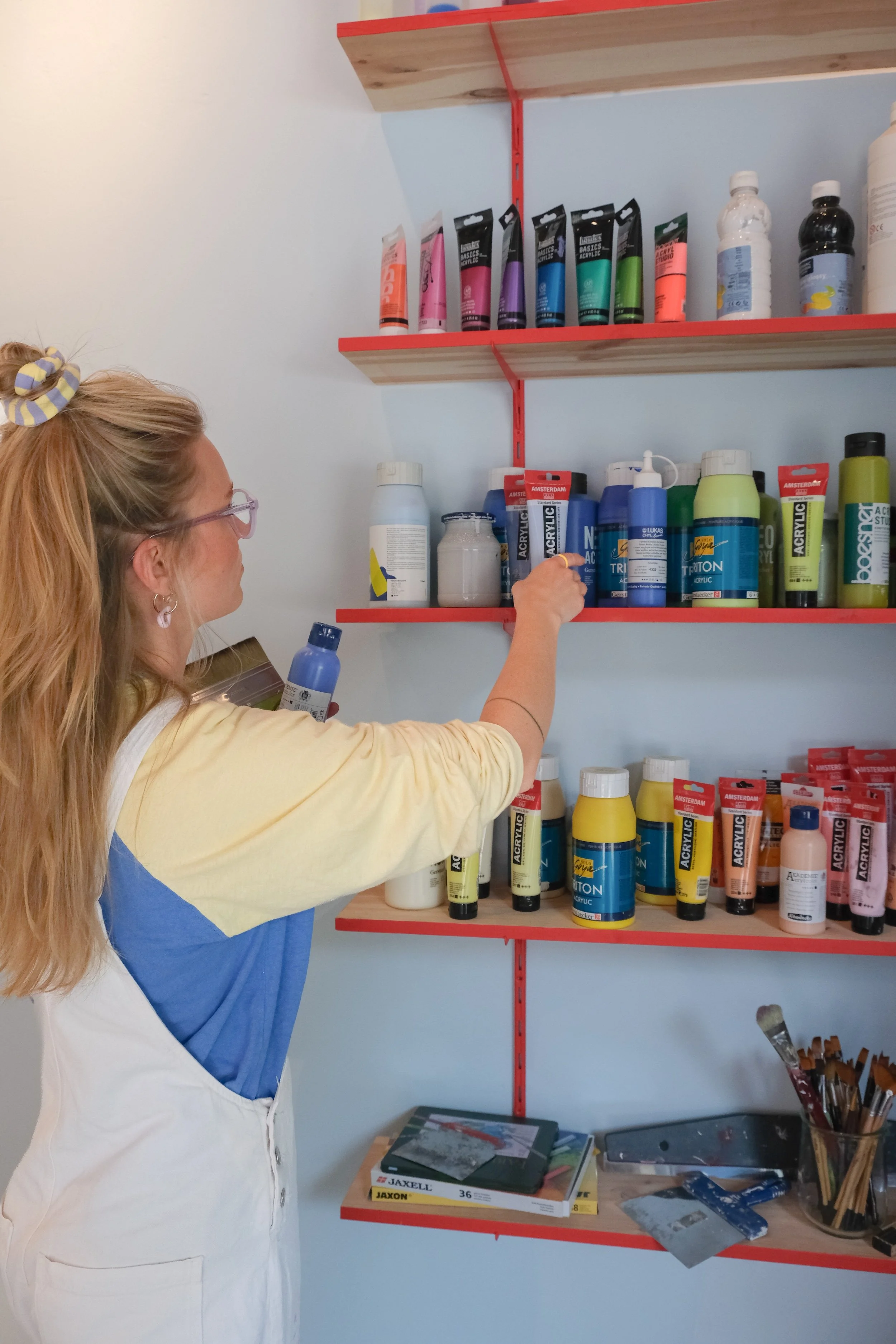 Eine Frau sortiert Acrylfarben in einer Bastel- oder Künstlerwerkstatt. Es gibt Regale mit verschiedenen Farbtuben, Gläser und Bastelmaterialien auf einem Tisch.