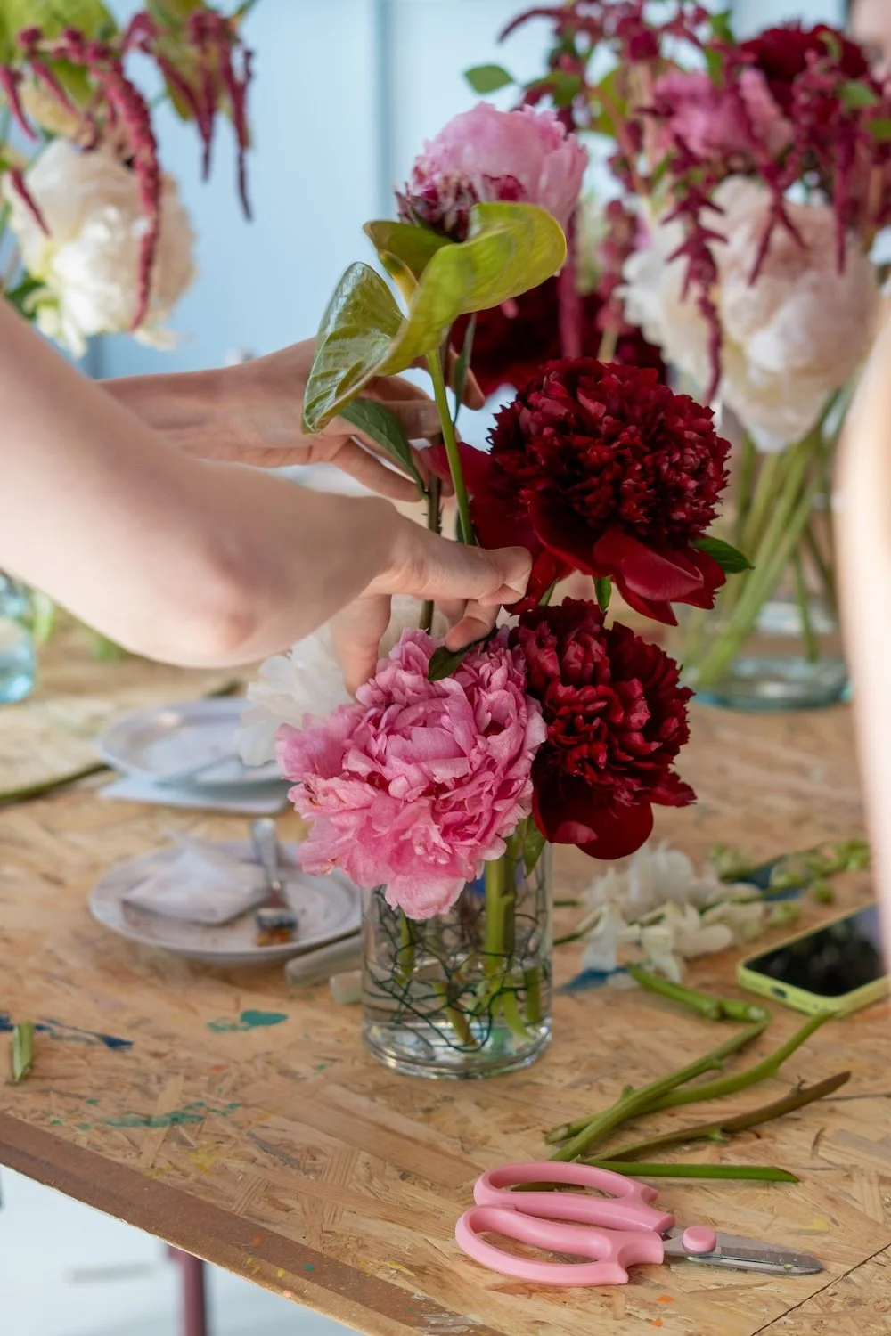 Person arrangiert eine bunte Blumenstrauß, bestehend aus pinken, roten und weißen Blumen in einer Glasvase, auf einem Holztisch mit Gartenwerkzeug und Geschirr.