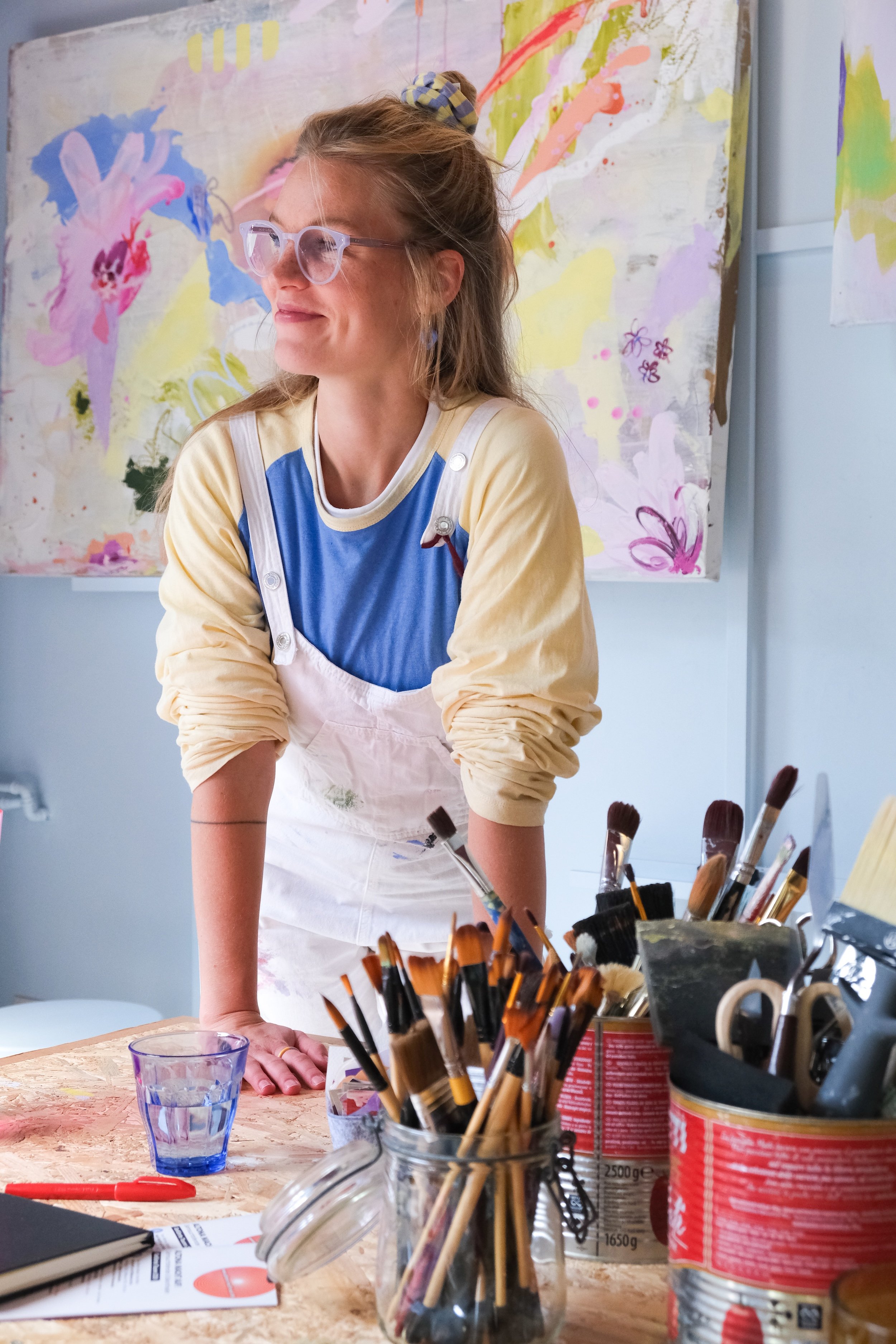 Eine Frau mit Brille und Haarband beim Malen in einem Atelier. Im Hintergrund bunte, abstrakte Gemälde, und Pinsel in Dosen auf einem Arbeitstisch.
