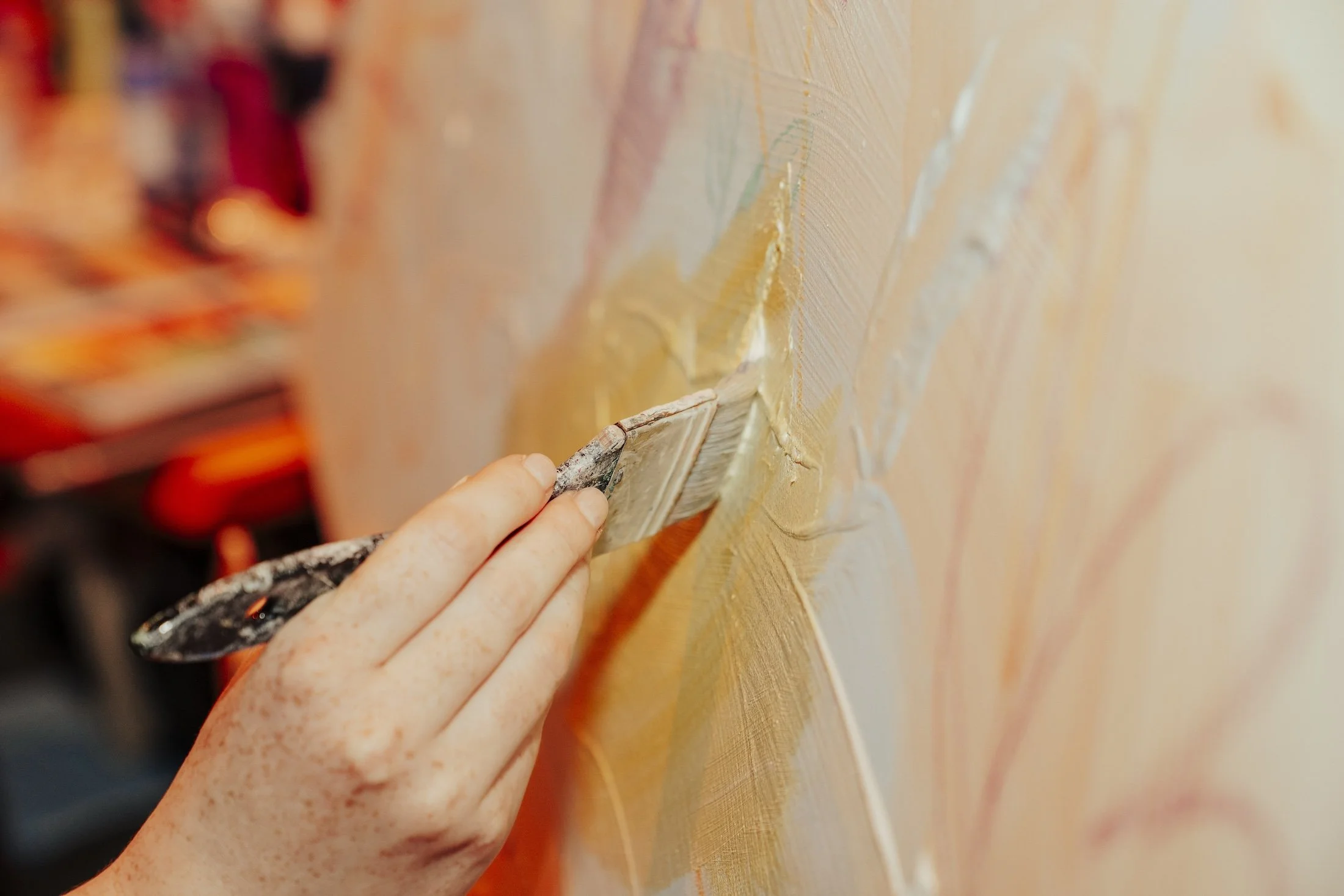 Hand hält Spachtel, der Farbe oder Gips auf eine Leinwand oder Wand aufträgt, im Hintergrund unscharfe Farben und Pinsel.