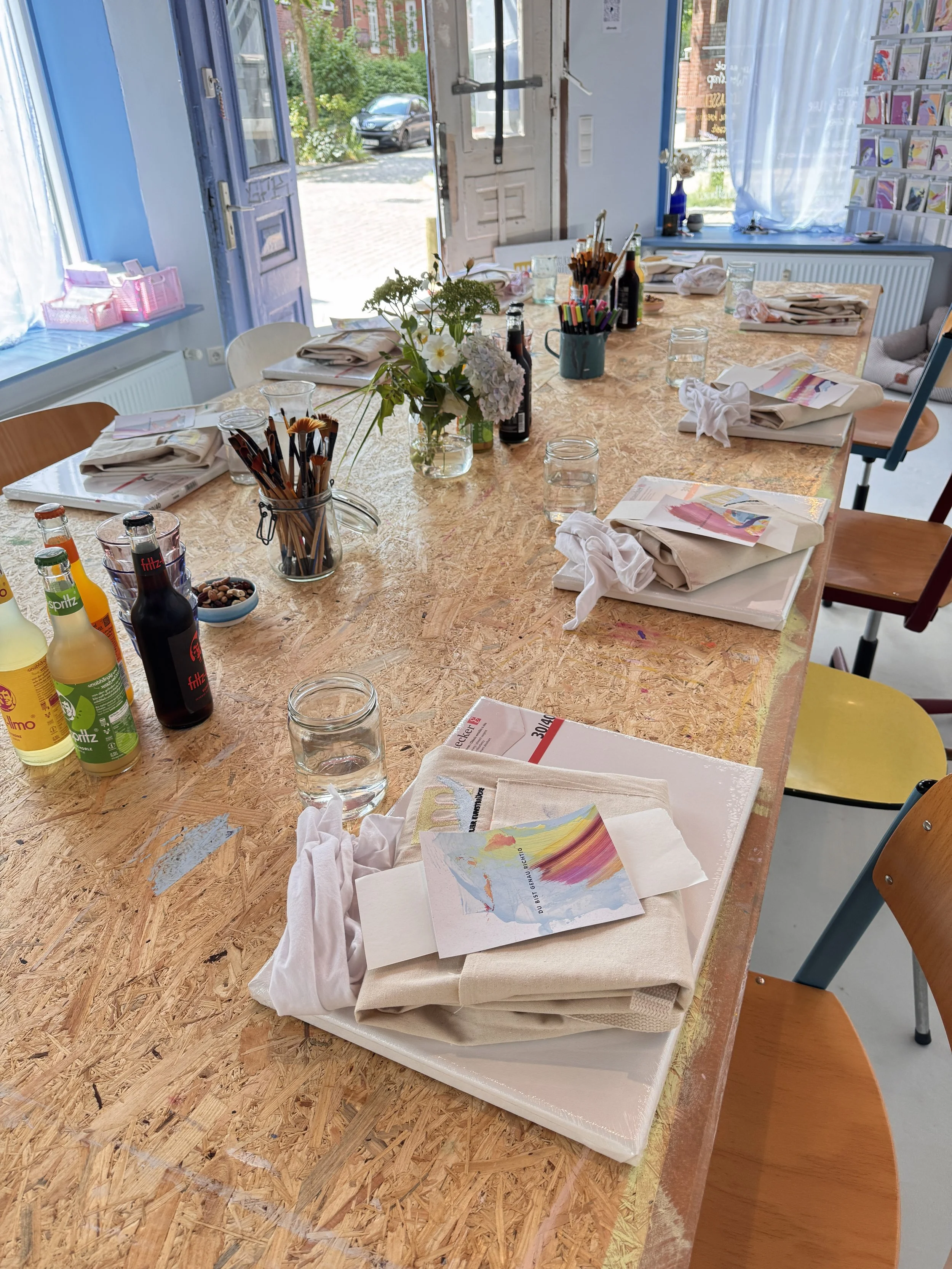 Ein heller Raum mit einer langen Holztisch, der für eine kreative Aktivität vorbereitet ist, mit Wasserflaschen, bunten Stiften, Papier, einer Blumenvase in der Mitte und Tüchern auf den Tellern. Im Hintergrund sind Fenster mit Blick nach draußen, Re