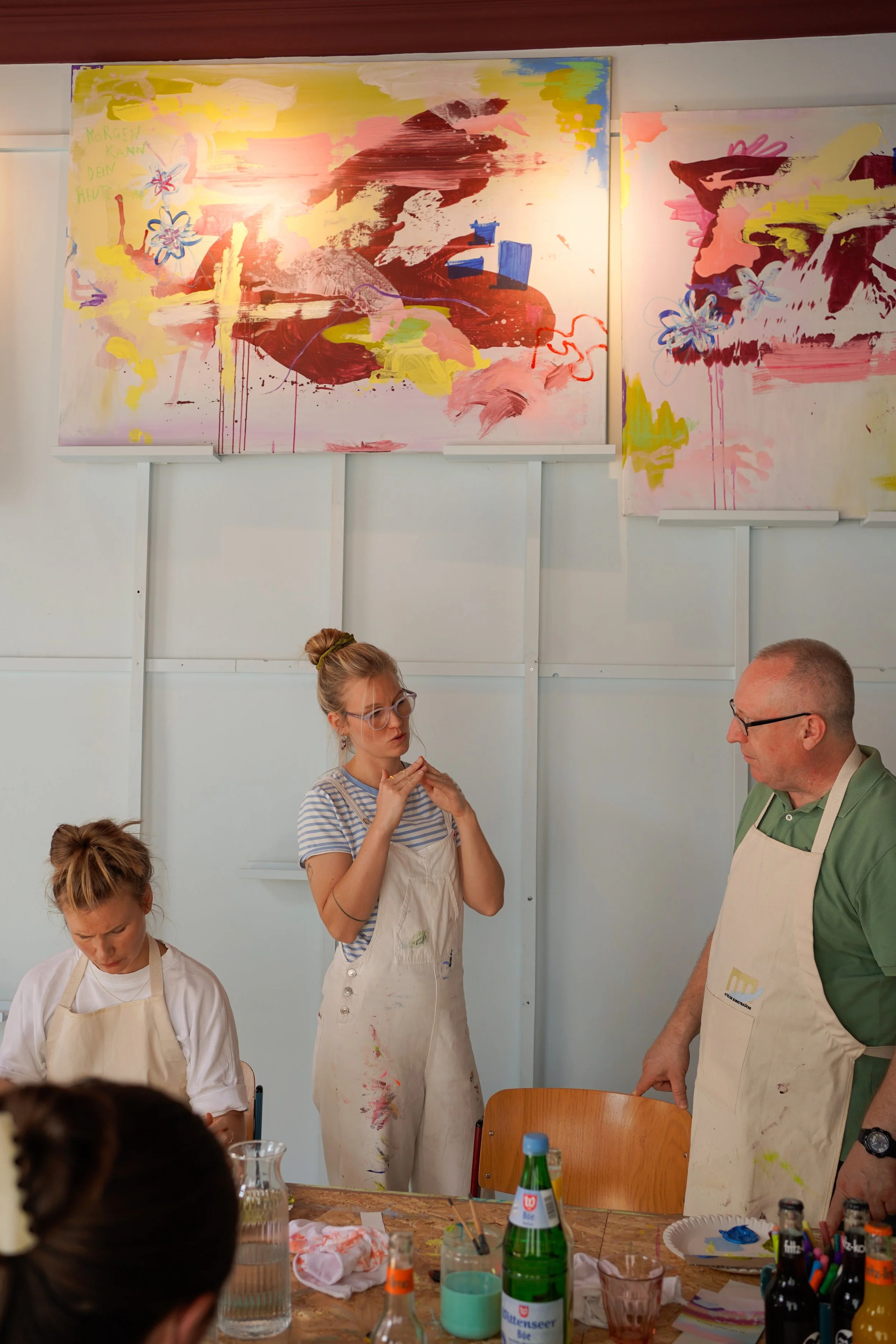 Menschen in einem Raum, die an einem Kunstprojekt arbeiten, mit mehreren bunten abstrakten Gemälden im Hintergrund, die auf der Wand hängen.